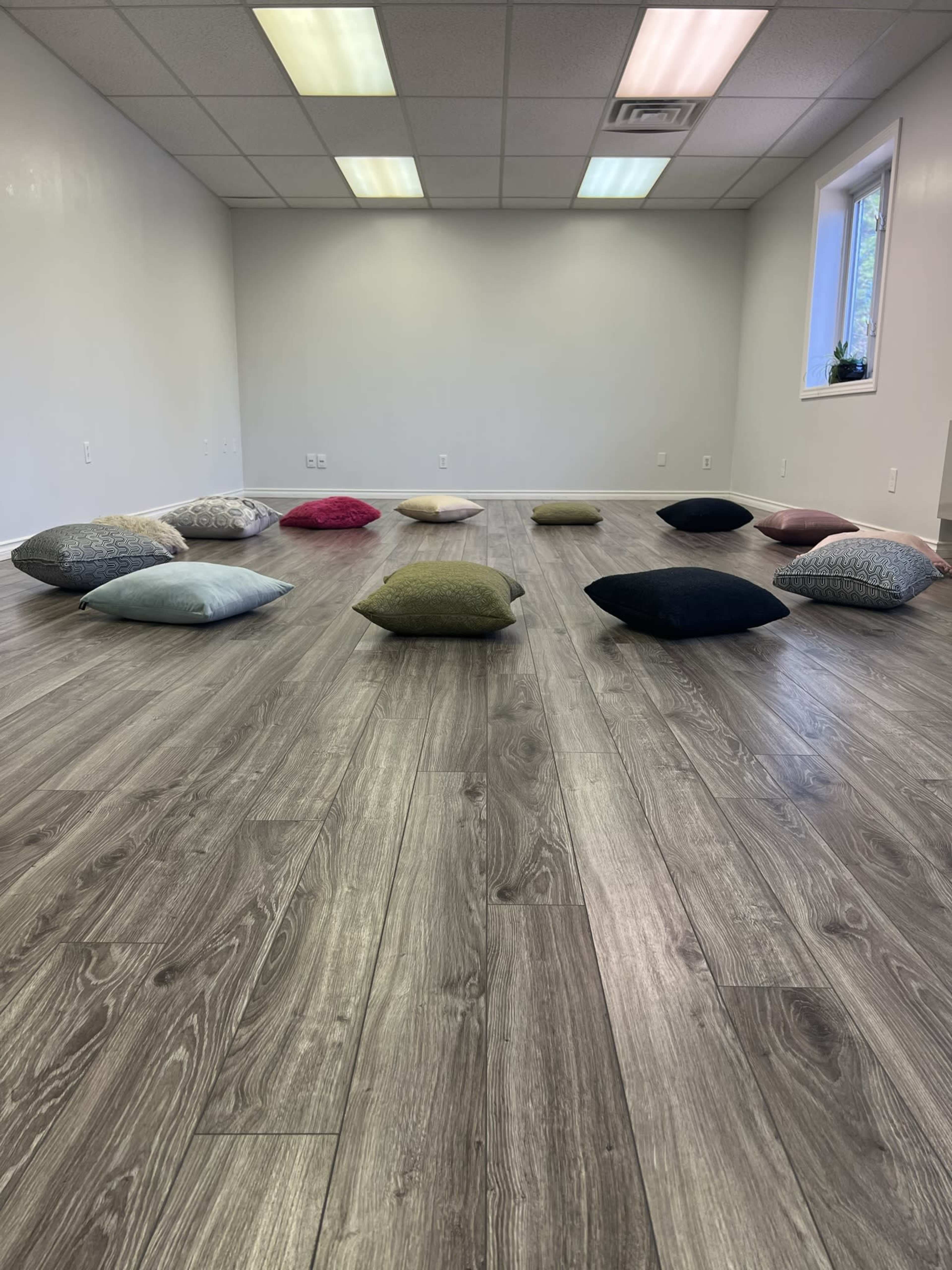The image shows a spacious, empty room with wooden flooring and various colored cushions arranged on the ground.