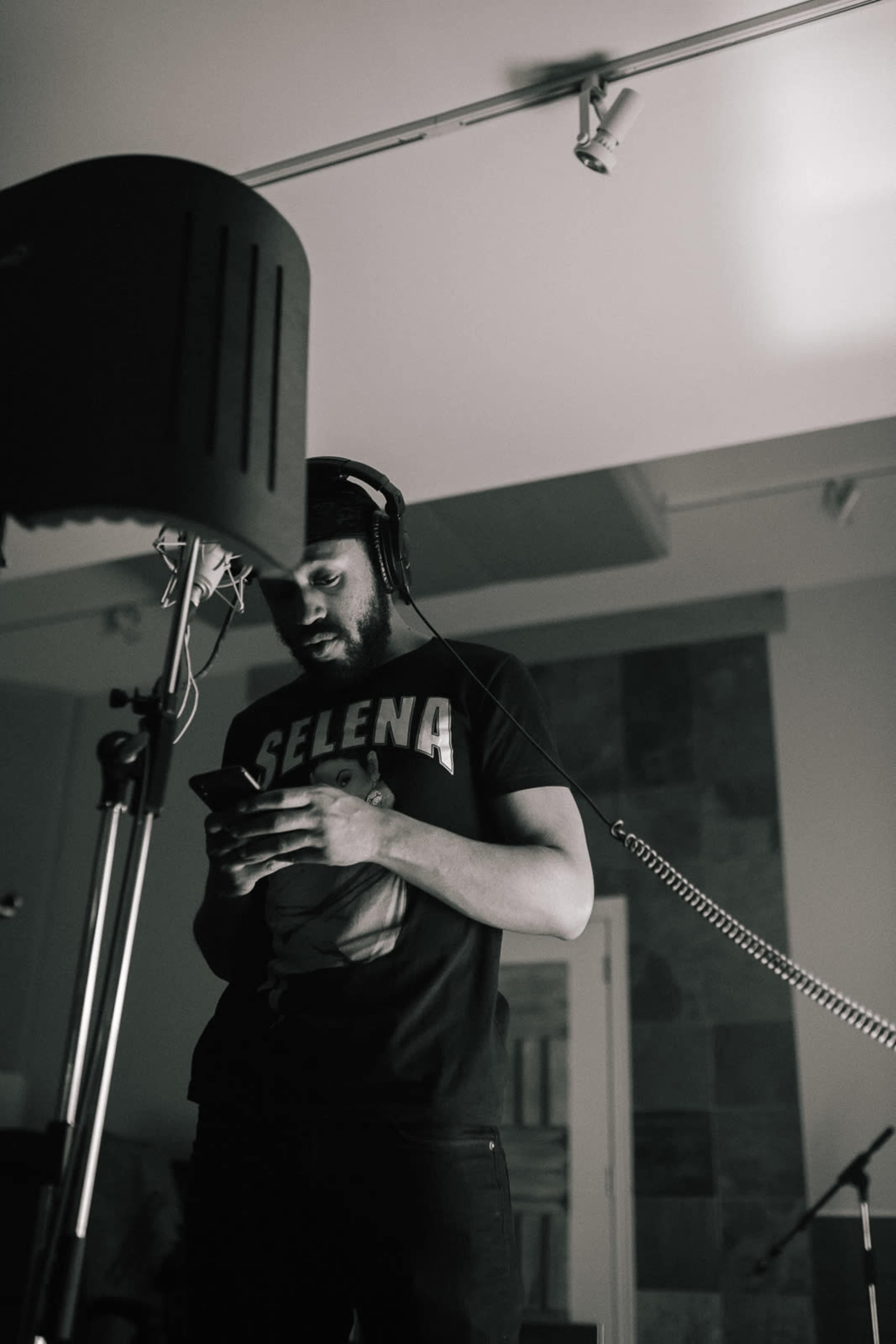 A man wearing a Selena t-shirt stands in a recording studio, looking at his phone while near a microphone.