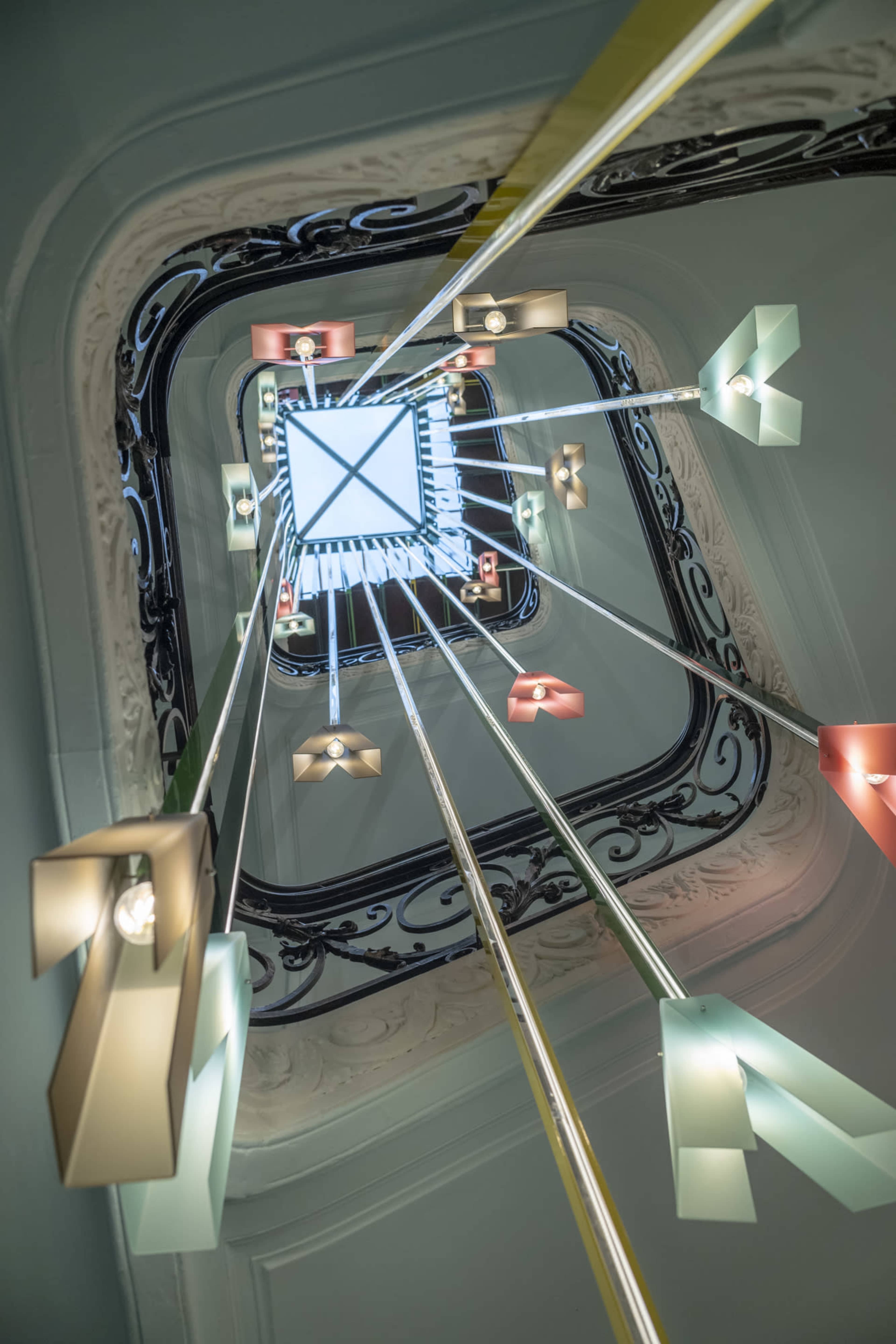 The image shows a spiral staircase with colorful hanging light fixtures radiating from a central square opening.