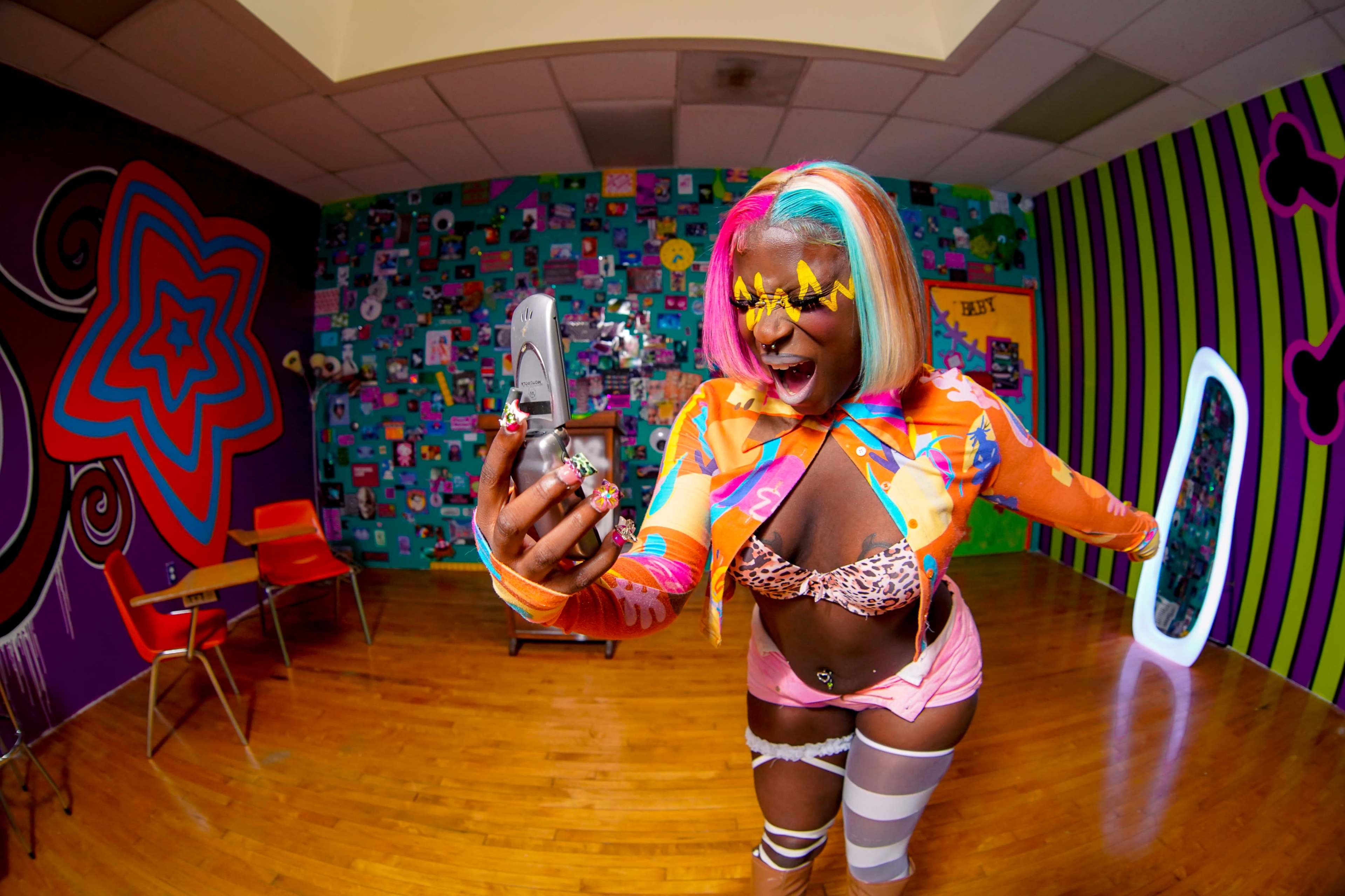 A person with colorful hair and makeup takes a selfie in a vibrantly decorated room filled with various patterns and colors.