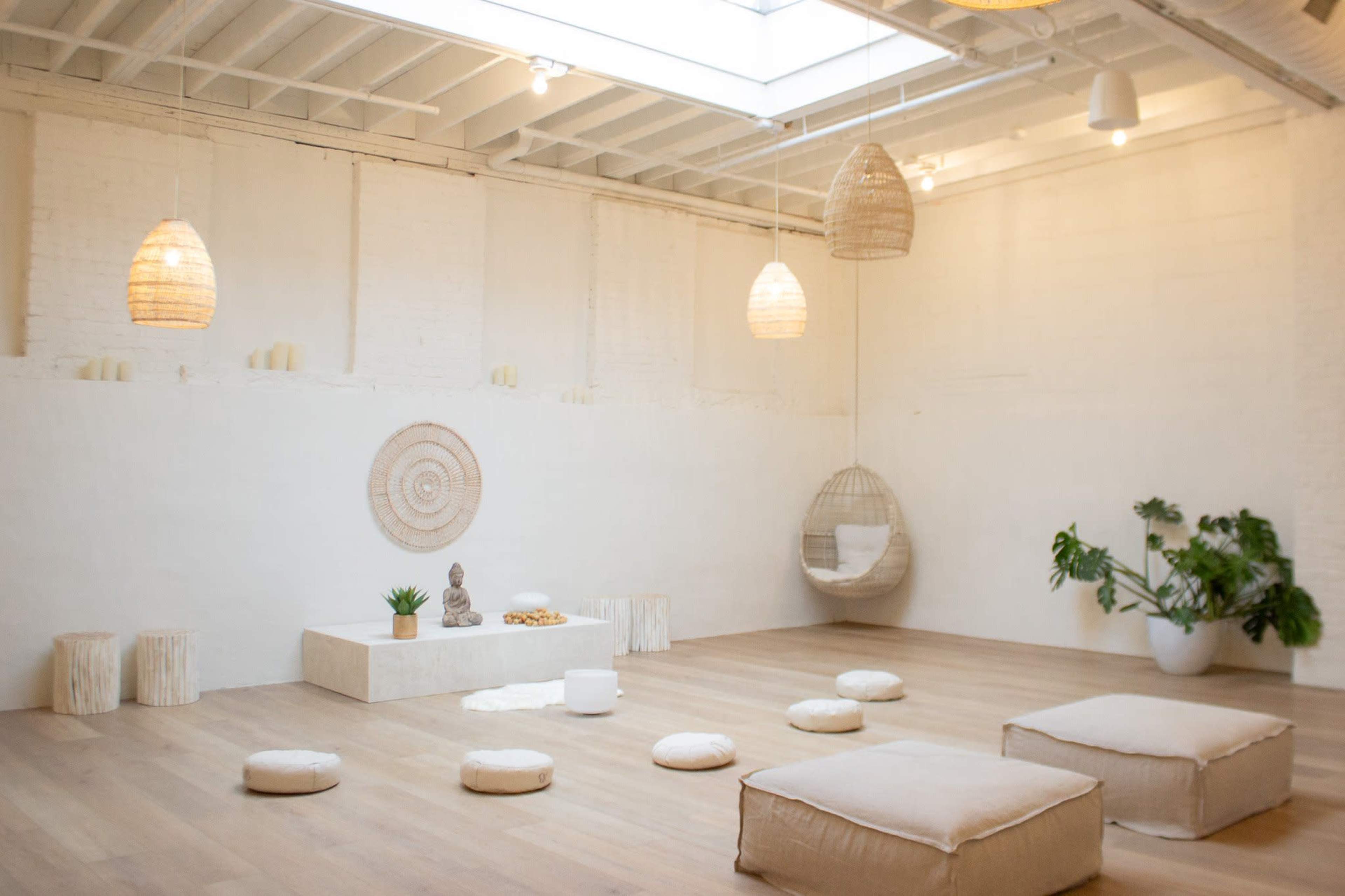 A minimalist meditation space with soft seating, a decorative wall hanging, a small statue, and plants, illuminated by natural light from a skylight.