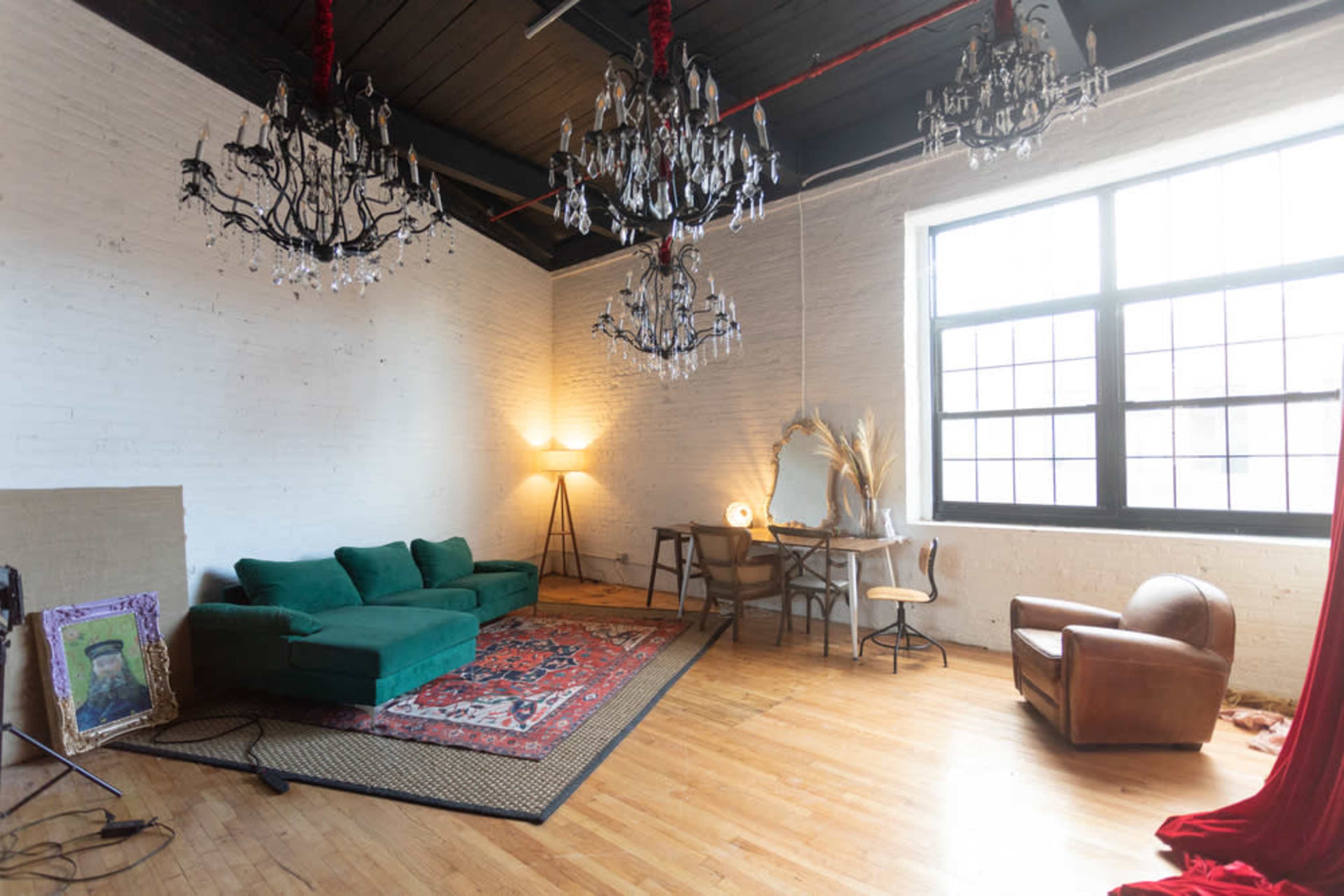 The image depicts a spacious room with exposed brick walls, two large chandeliers, a green sofa, a wooden desk and chair, a vintage mirror, and a brown armchair, all arranged over a decorative rug.