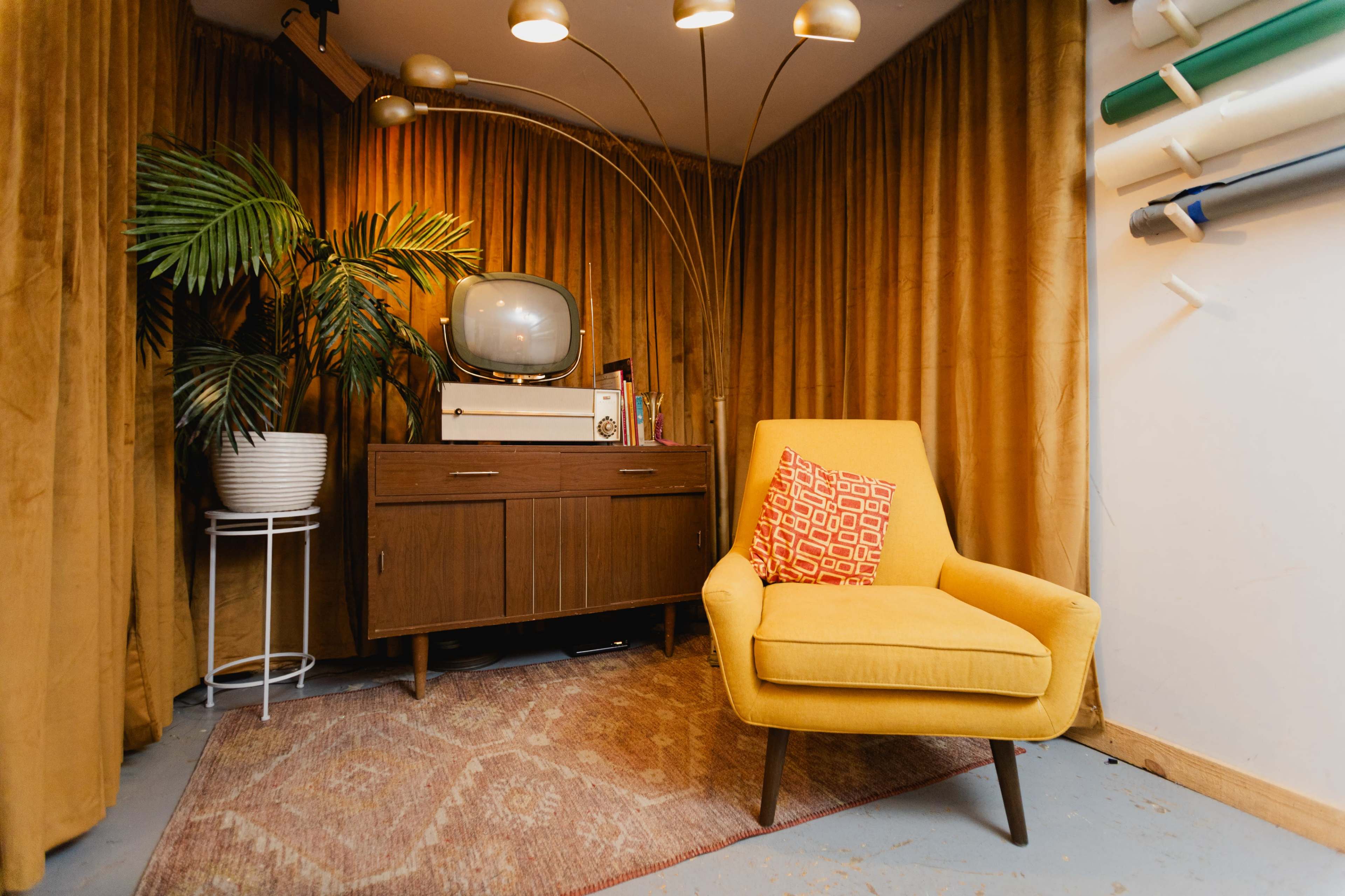 The image features a cozy corner of a room with a vintage television on a wooden cabinet, a yellow armchair, a decorative pillow, a potted plant, and a backdrop of gold curtains.