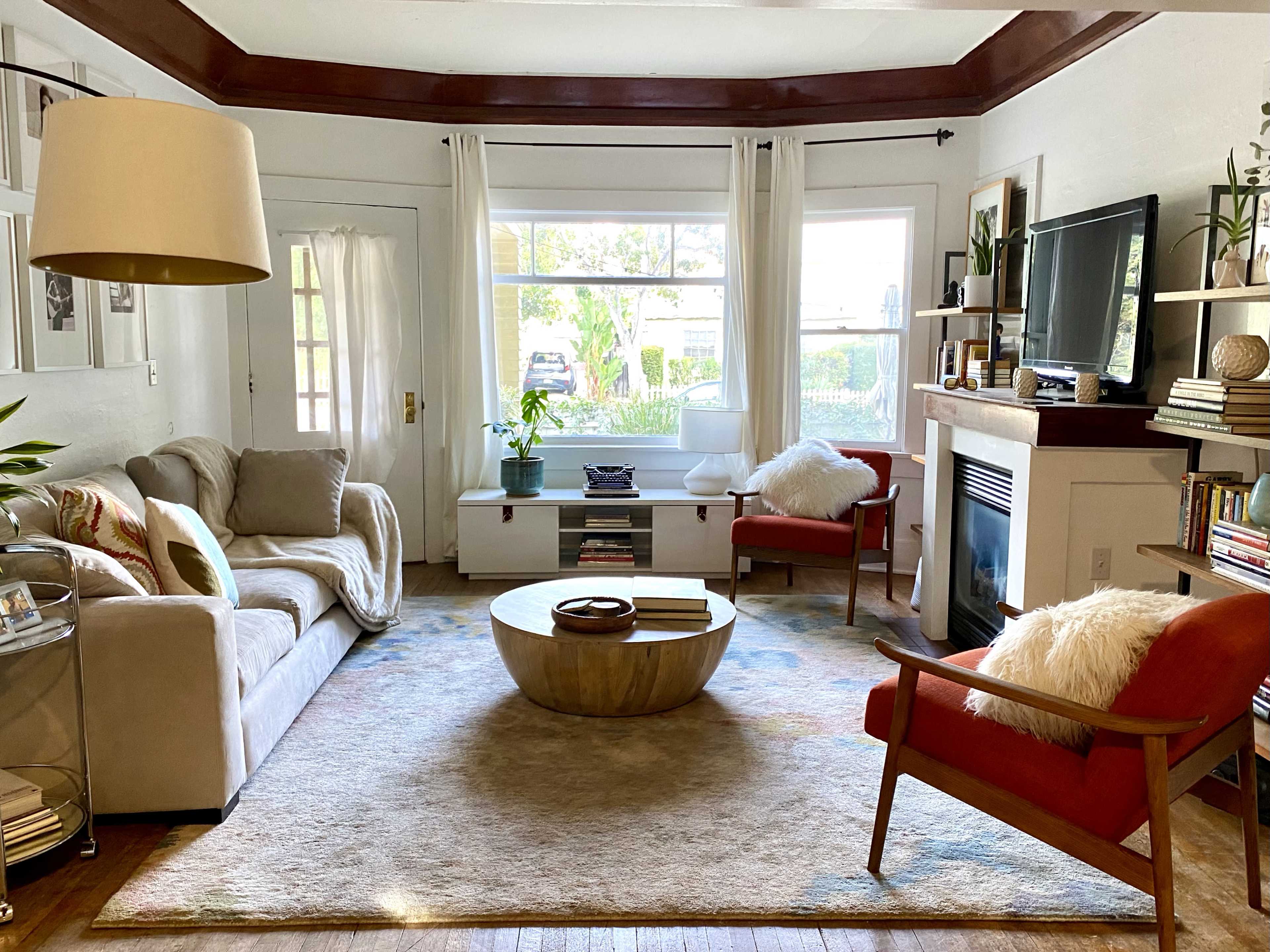 The image shows a cozy living room with a light gray sofa, two orange chairs, a round wooden coffee table, and large windows that provide natural light.
