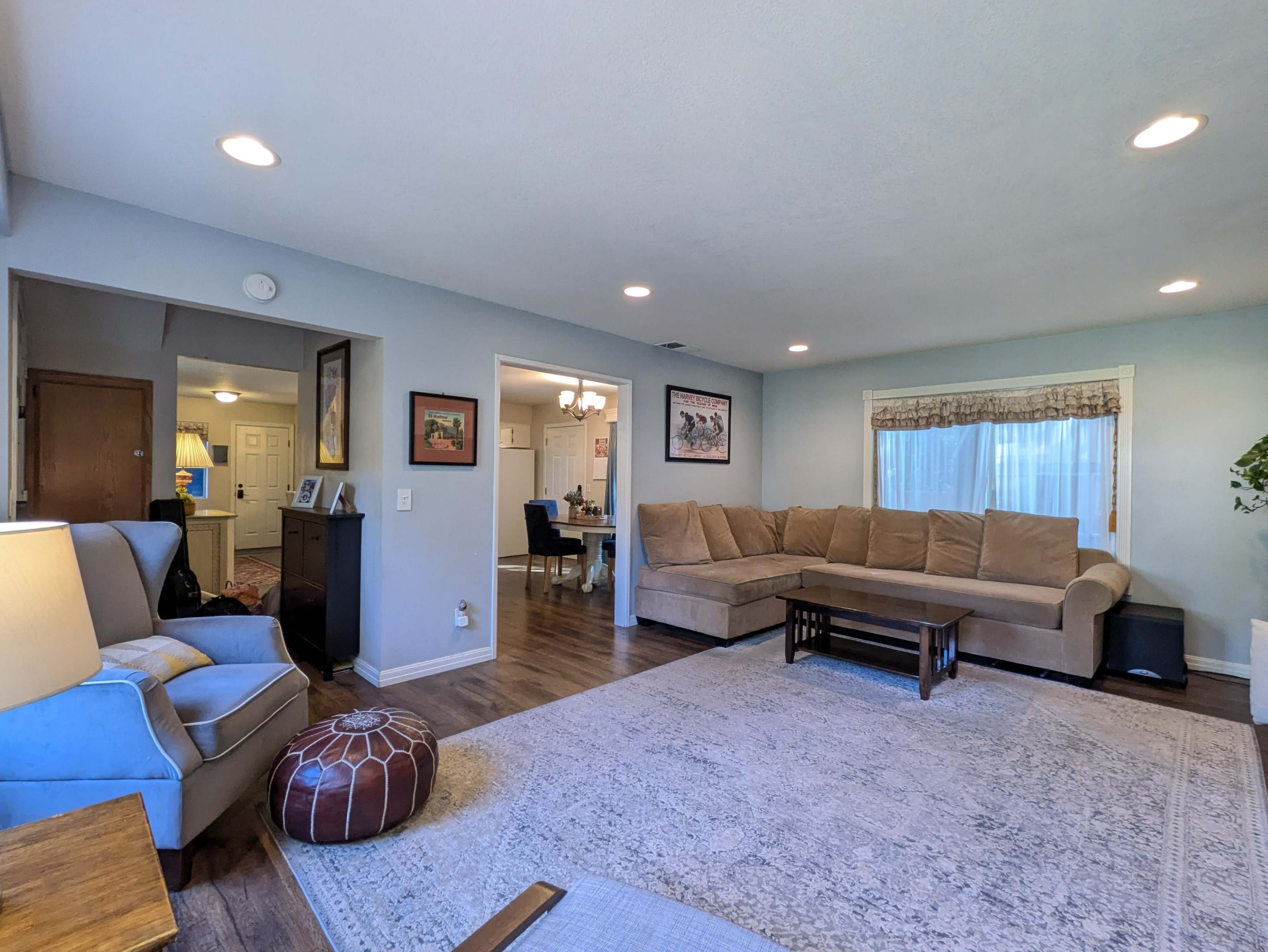 The image shows a spacious living room with a large beige sectional sofa, a coffee table, and a decorative rug, set against light blue walls and wooden flooring.