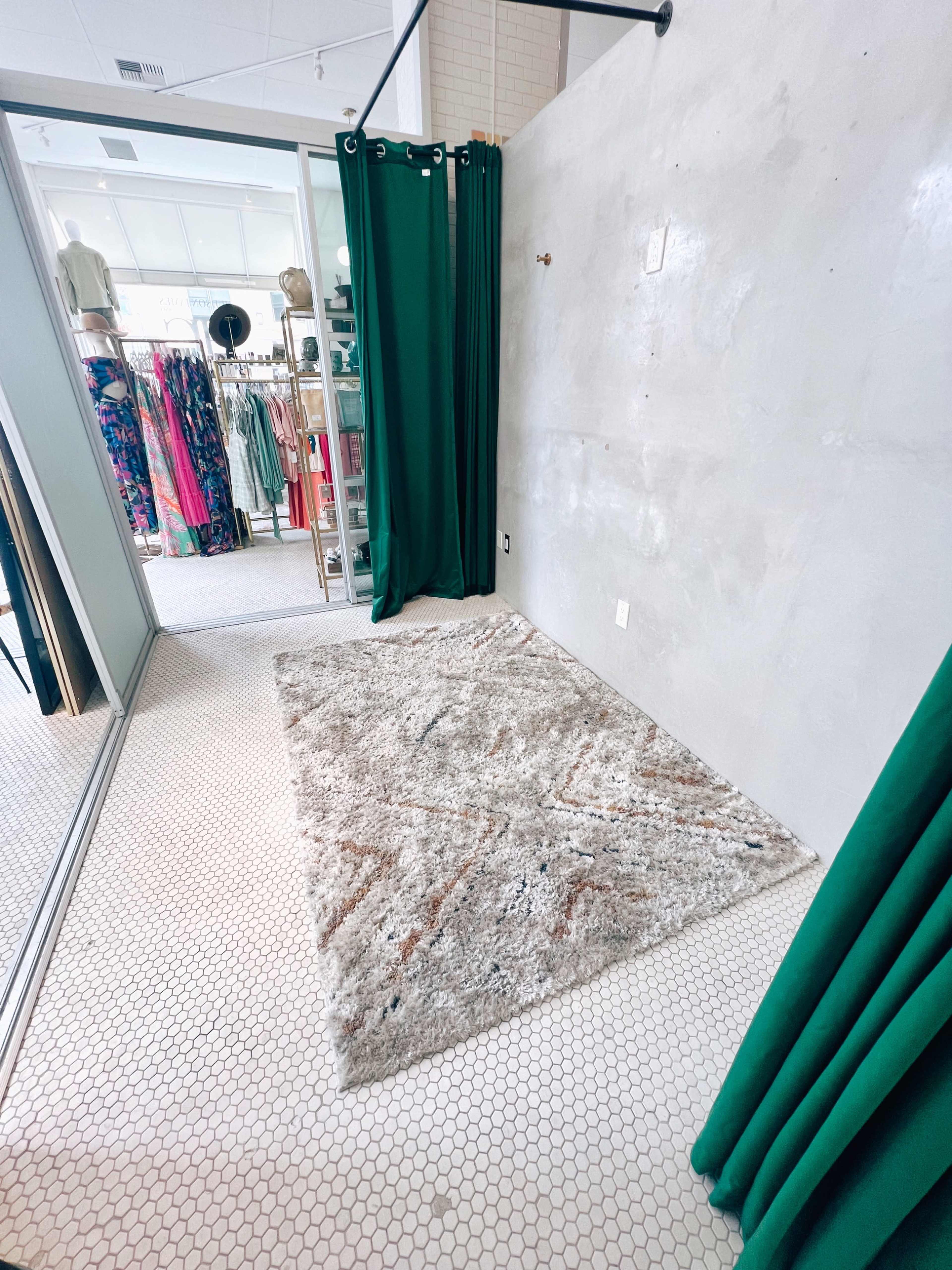 A dressing room with a textured gray wall, a patterned rug on a hexagonal tiled floor, and green curtains partially drawn.