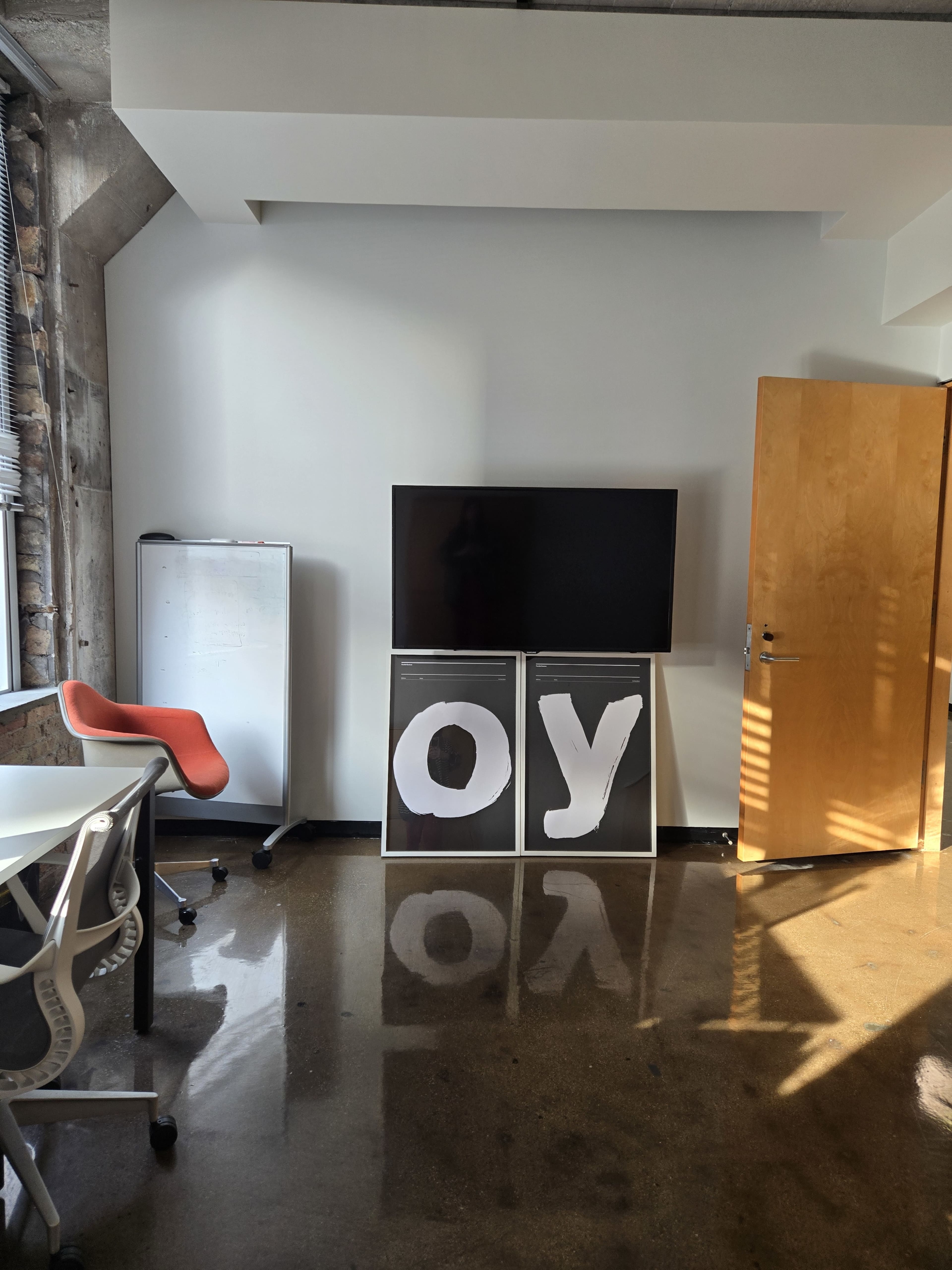 The image shows a modern office space with a large TV mounted on the wall, two framed pieces displaying the letters "O" and "Y," and a wooden door on the right side.