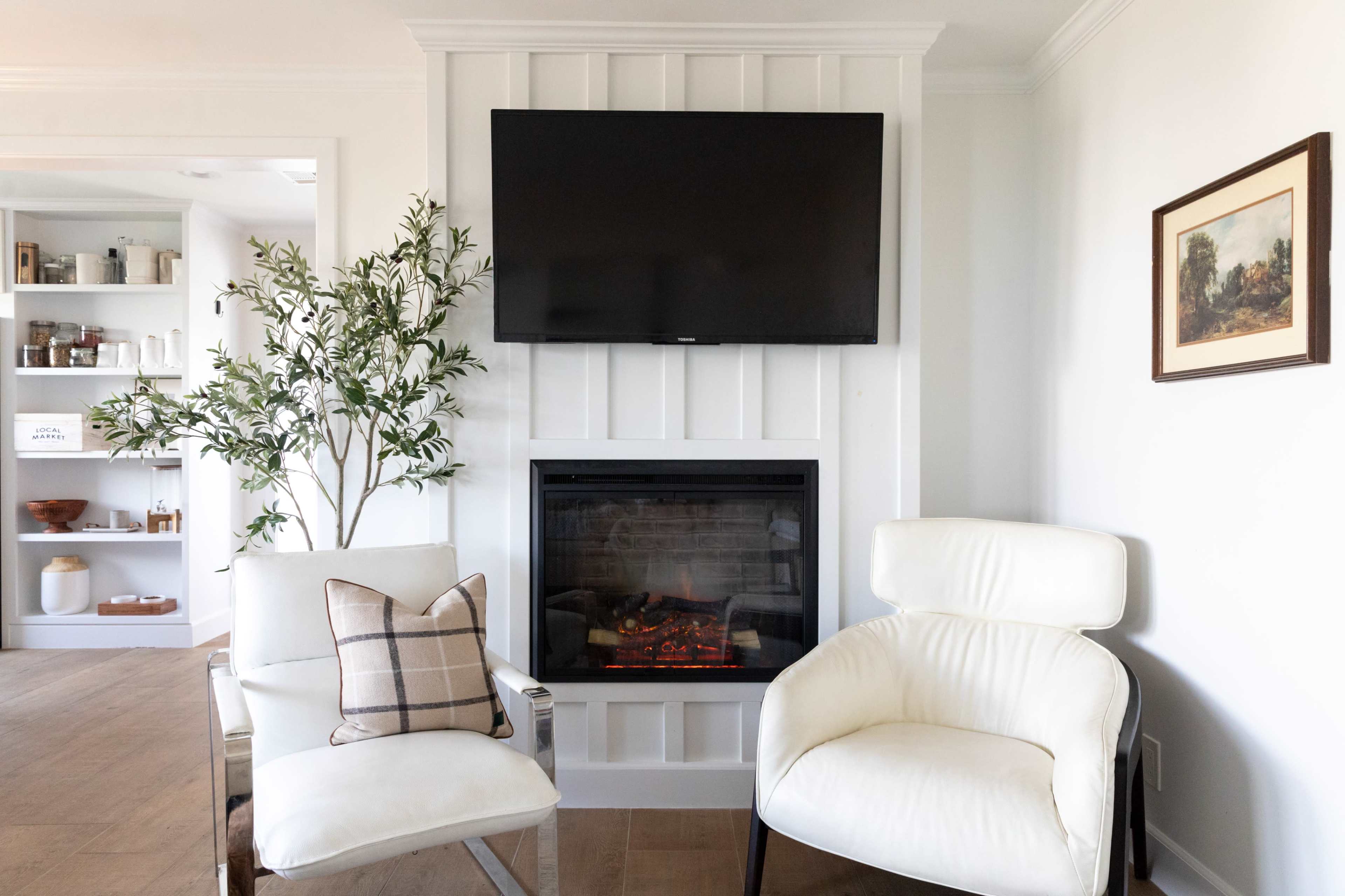 The image shows a living room with two white armchairs, a television mounted on a wall above a fireplace, and a decorative plant on the side.
