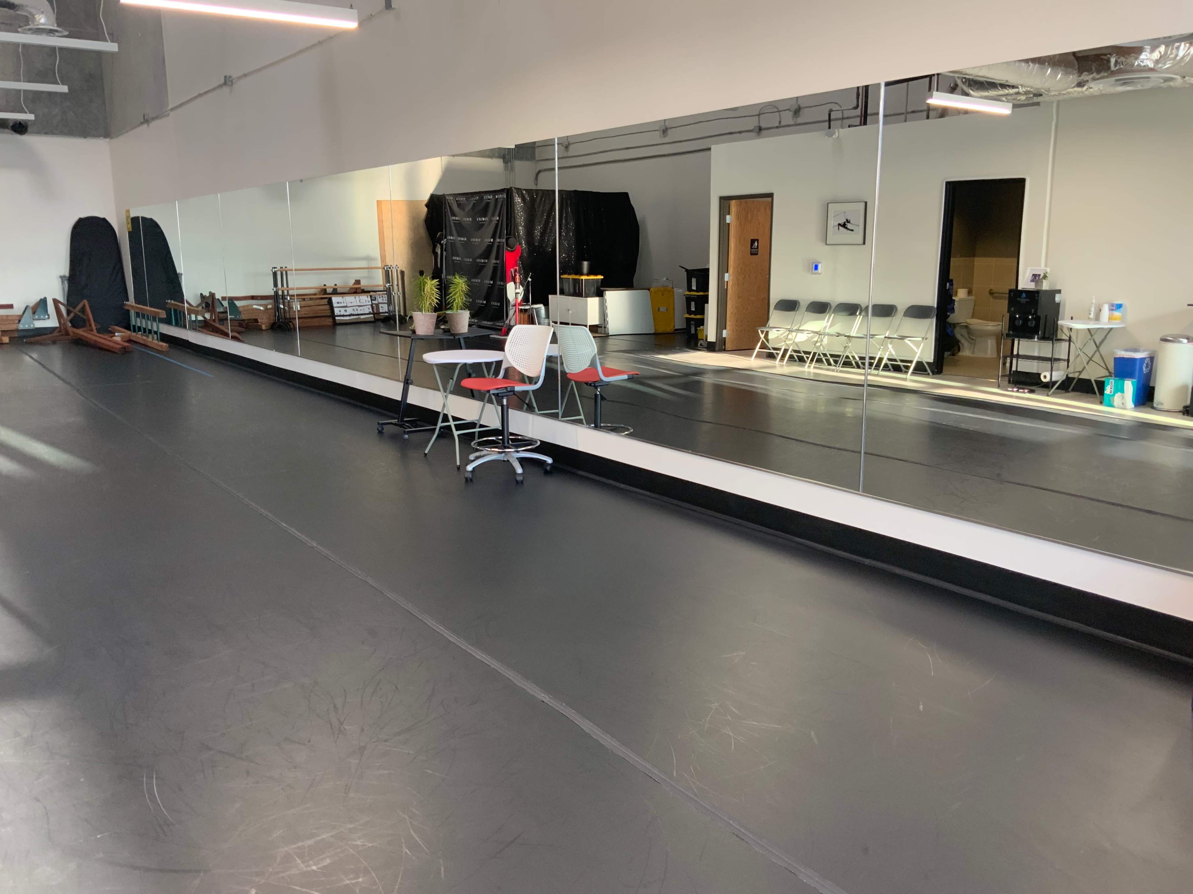 A dance studio features a mirrored wall, black flooring, and several chairs arranged around a small table.