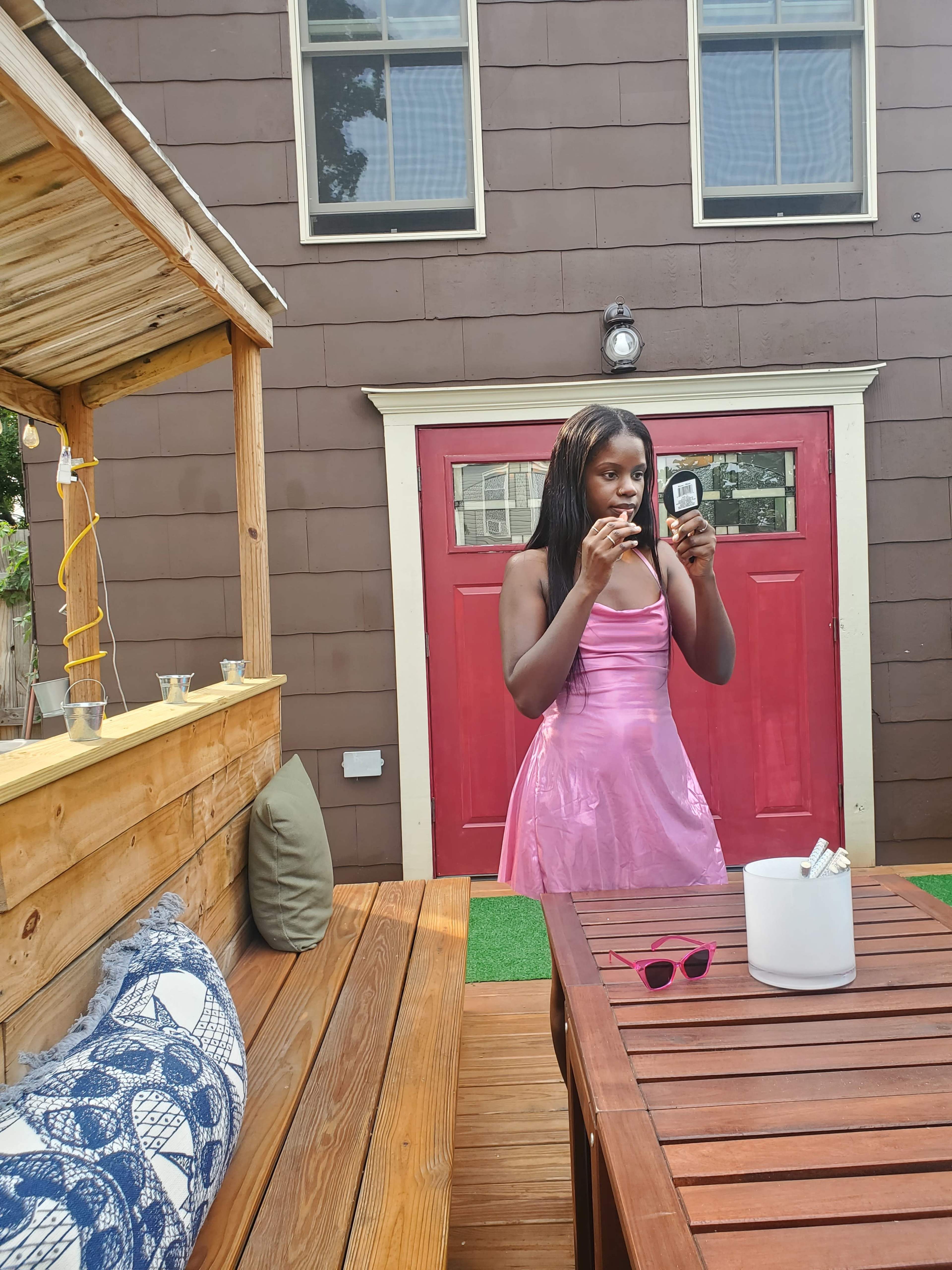 A person in a pink dress stands by a wooden table, applying lip gloss while a red door is visible in the background.