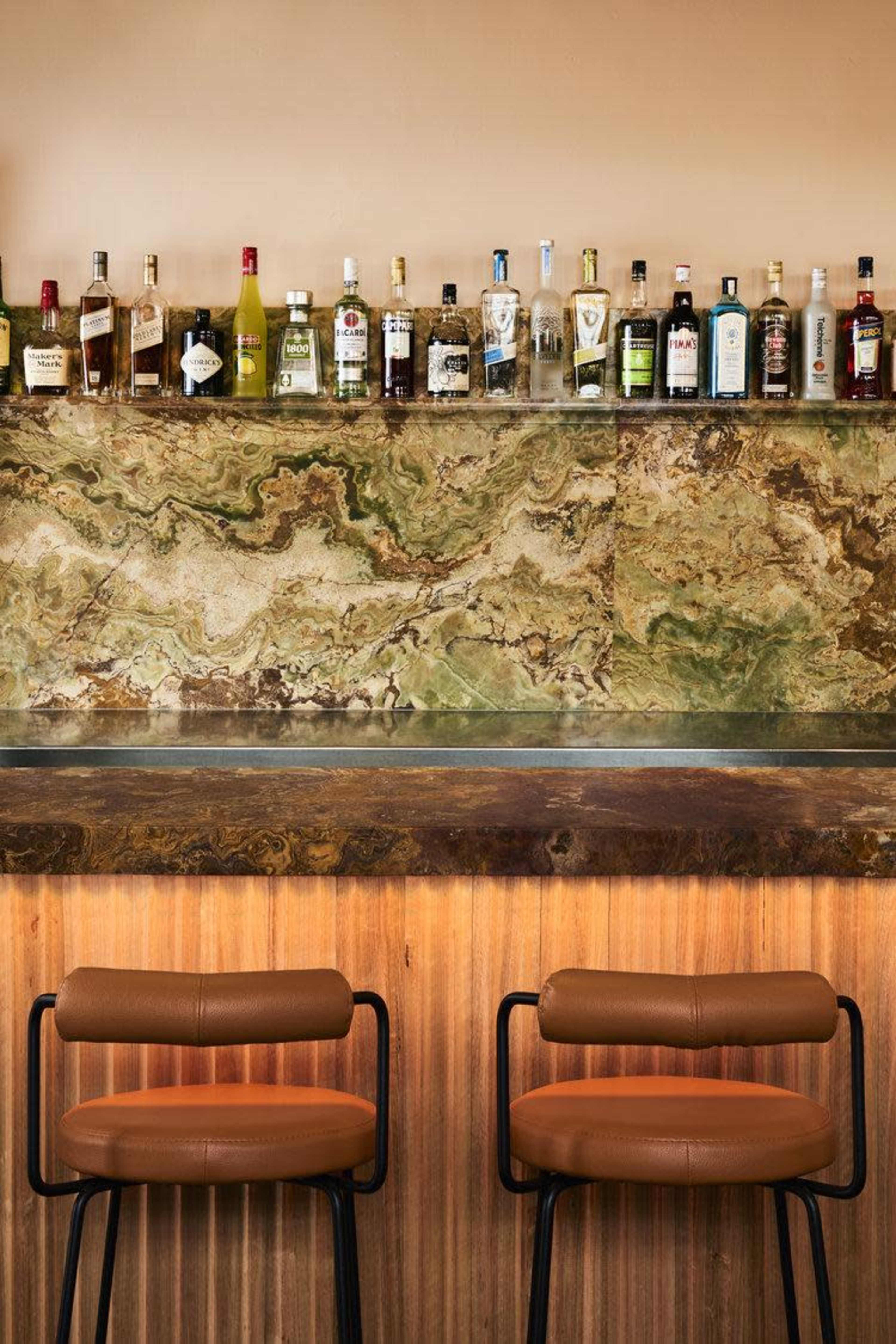 A bar with a selection of bottles displayed atop a green marble counter, accompanied by two brown leather stools in front.