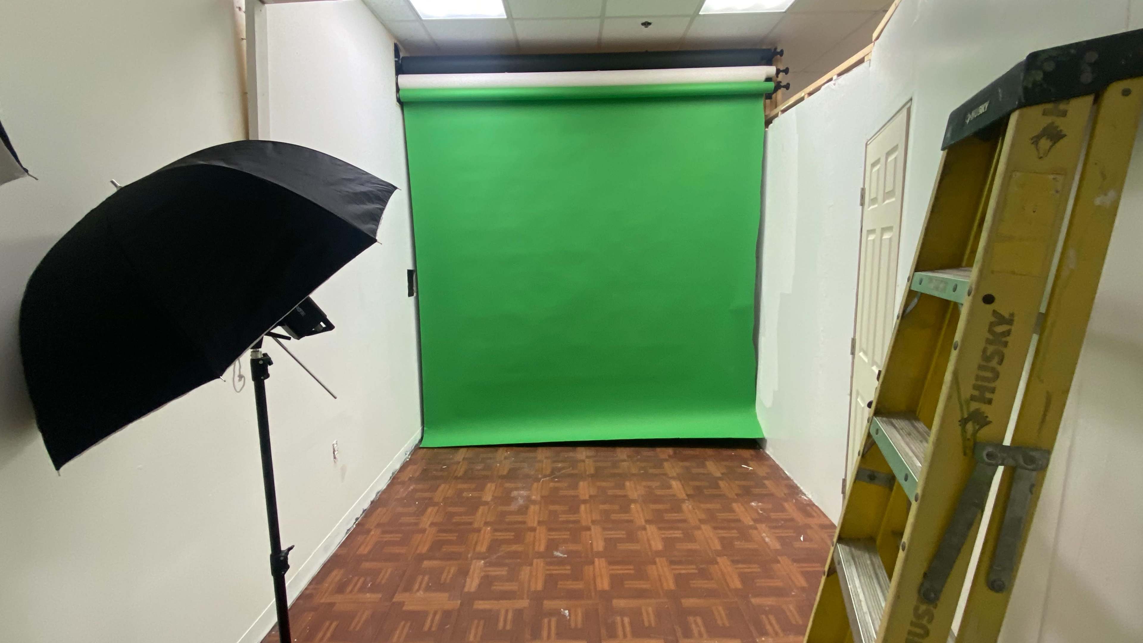A small room with a green screen backdrop, a black umbrella light, and a ladder positioned against the wall.