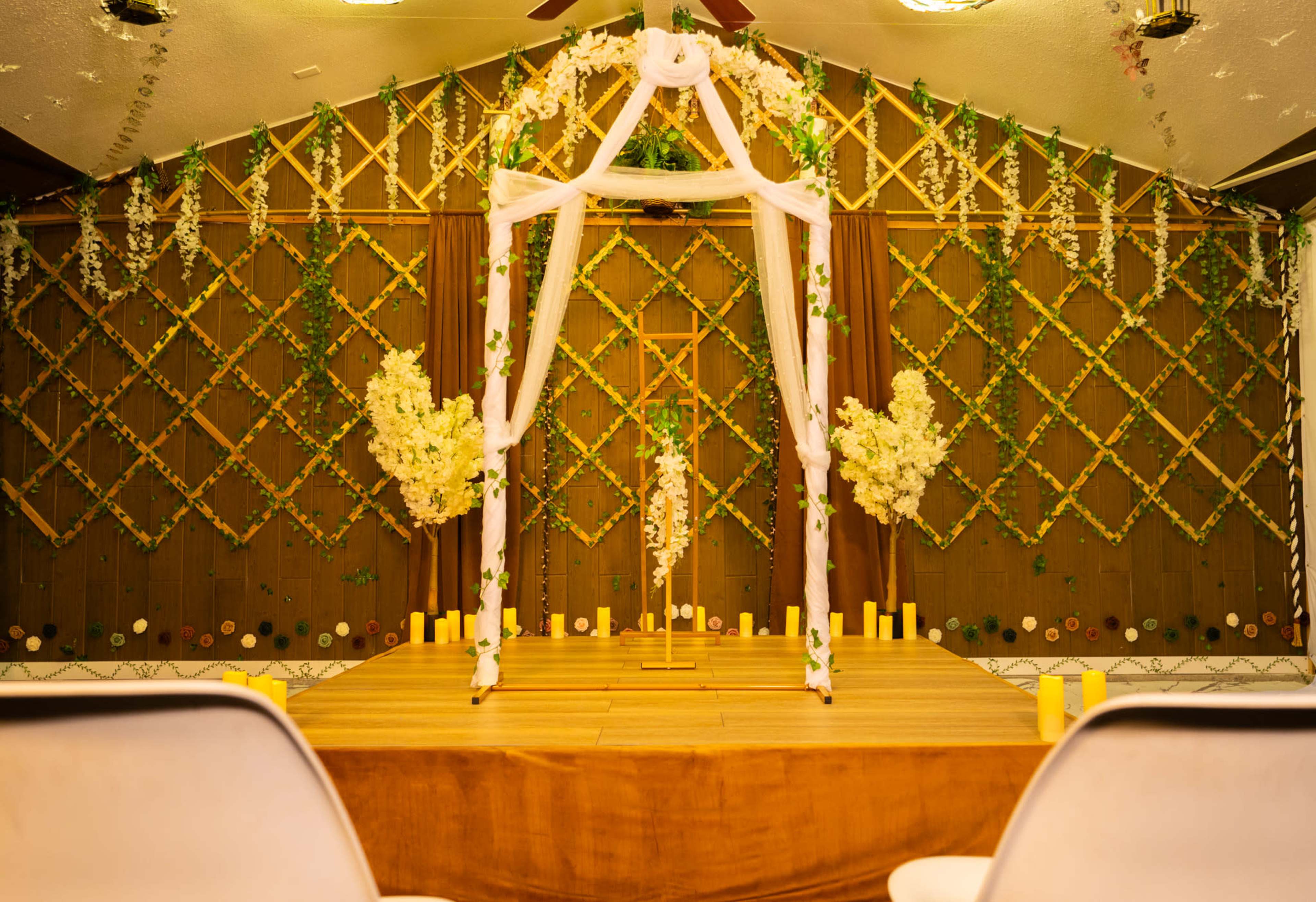 The image shows a decorated indoor space featuring a floral canopy in front of a wooden wall, with flowers and candles arranged on a stage.