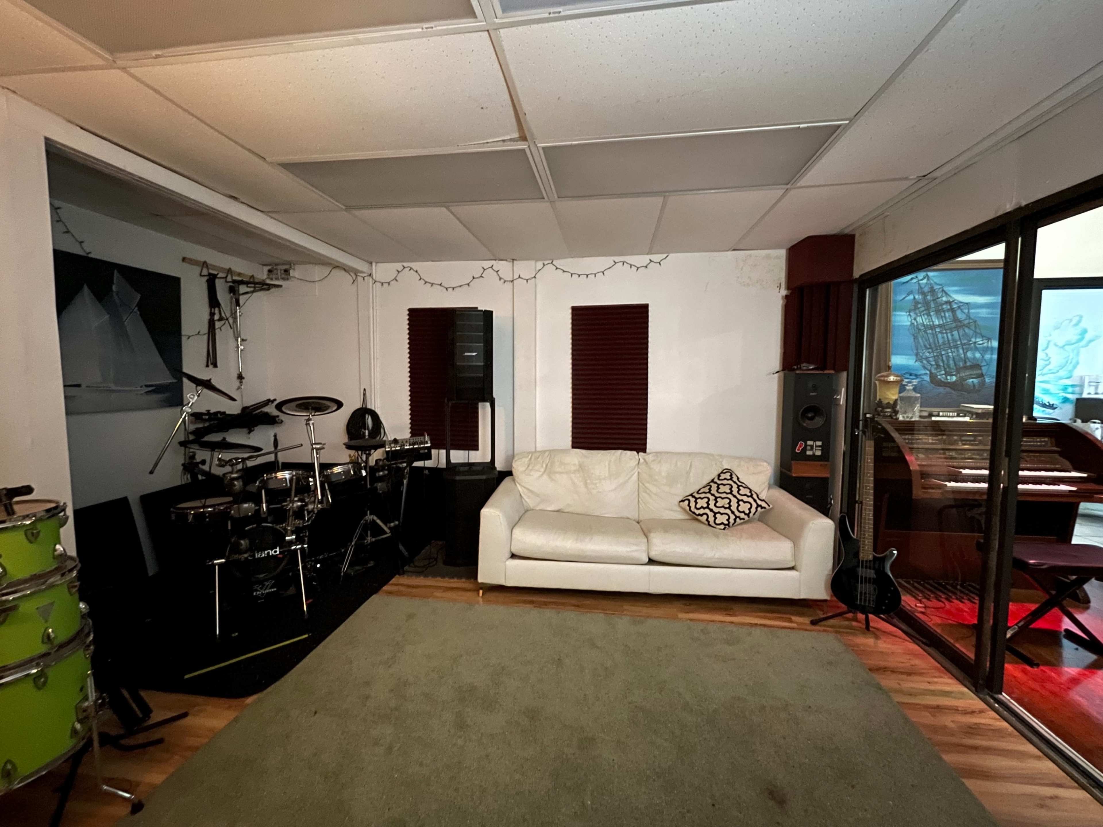 The room features a white couch facing a music setup with electronic drums, speakers, and a vintage keyboard, alongside a green area rug on wooden flooring.
