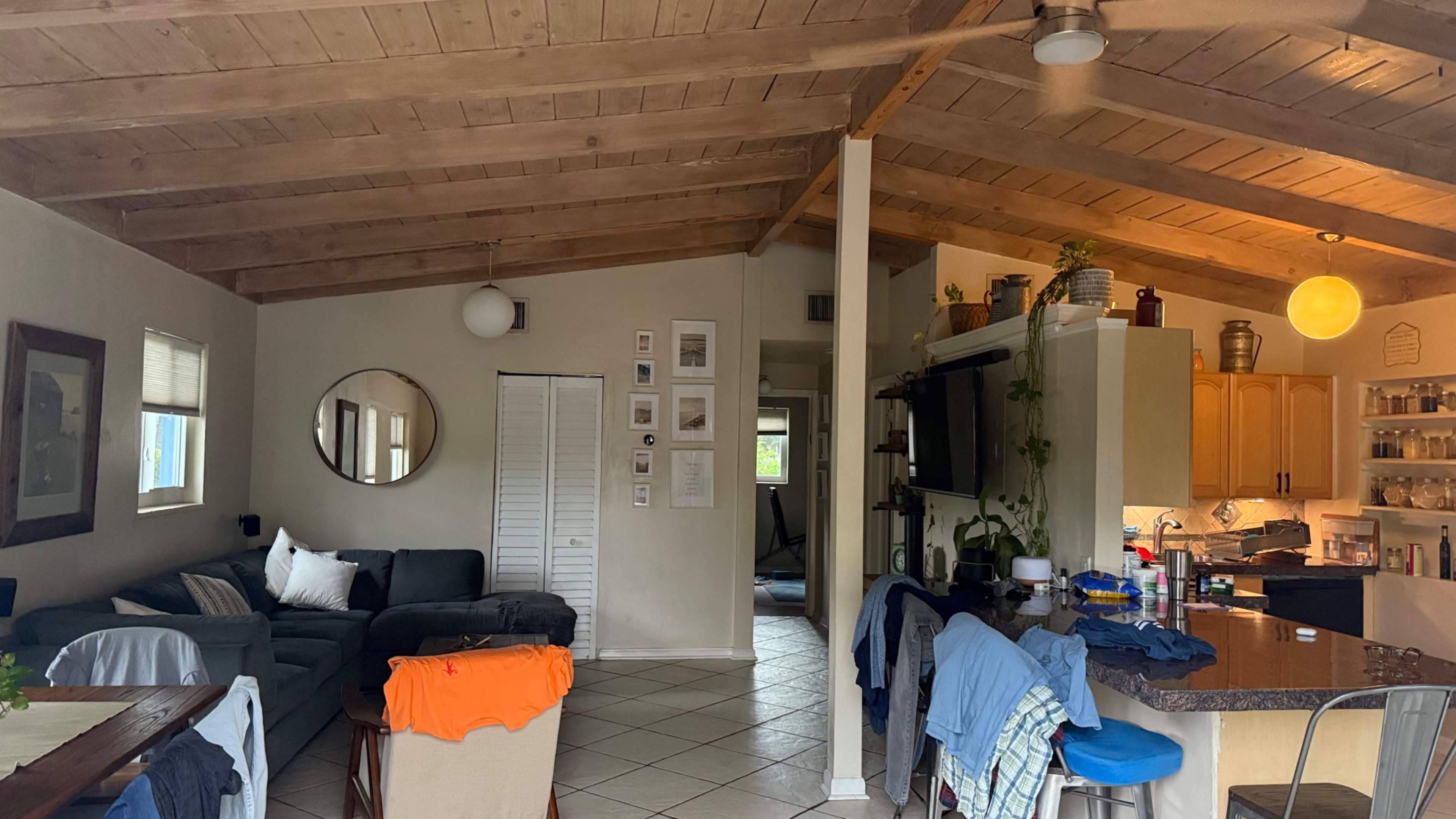 The image shows a living area featuring a gray sofa, wooden ceiling beams, and an open kitchen with a bar counter.