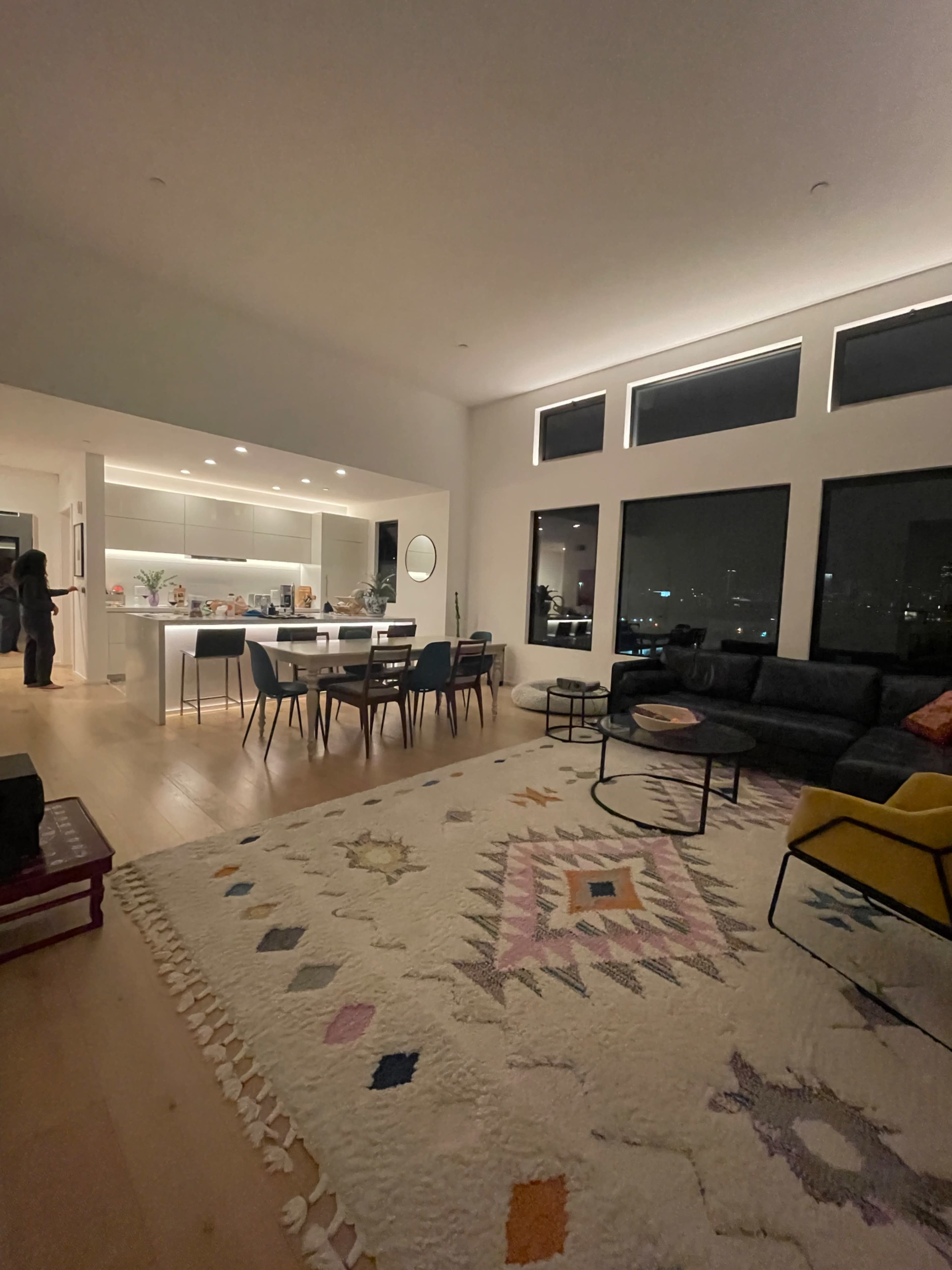 A modern living room features a large rug, a sectional sofa, a dining area with a table and chairs, and large windows overlooking a city view.