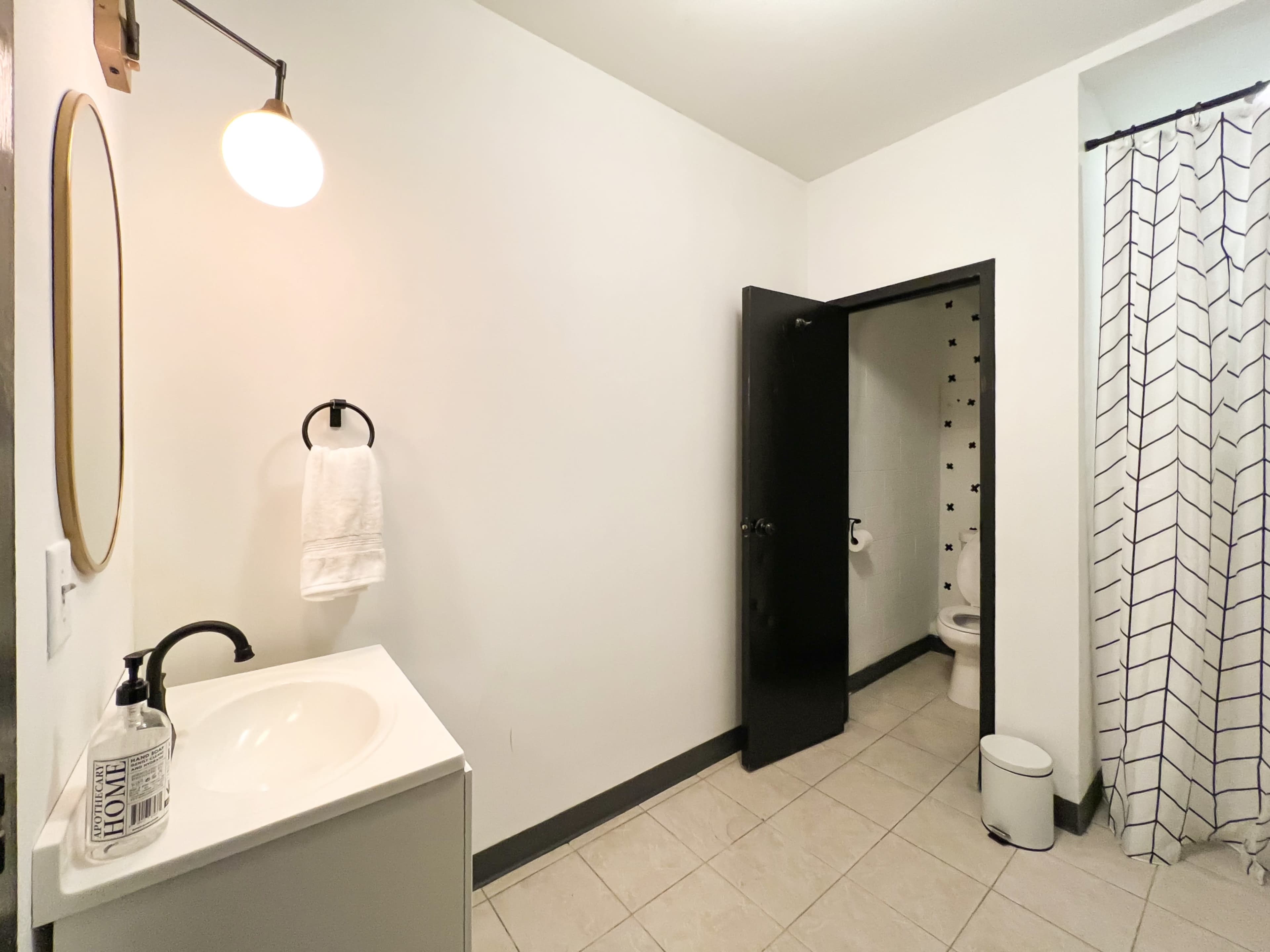 A well-lit bathroom features a sink with a mirror, a towel holder, and an open door leading to a toilet area.