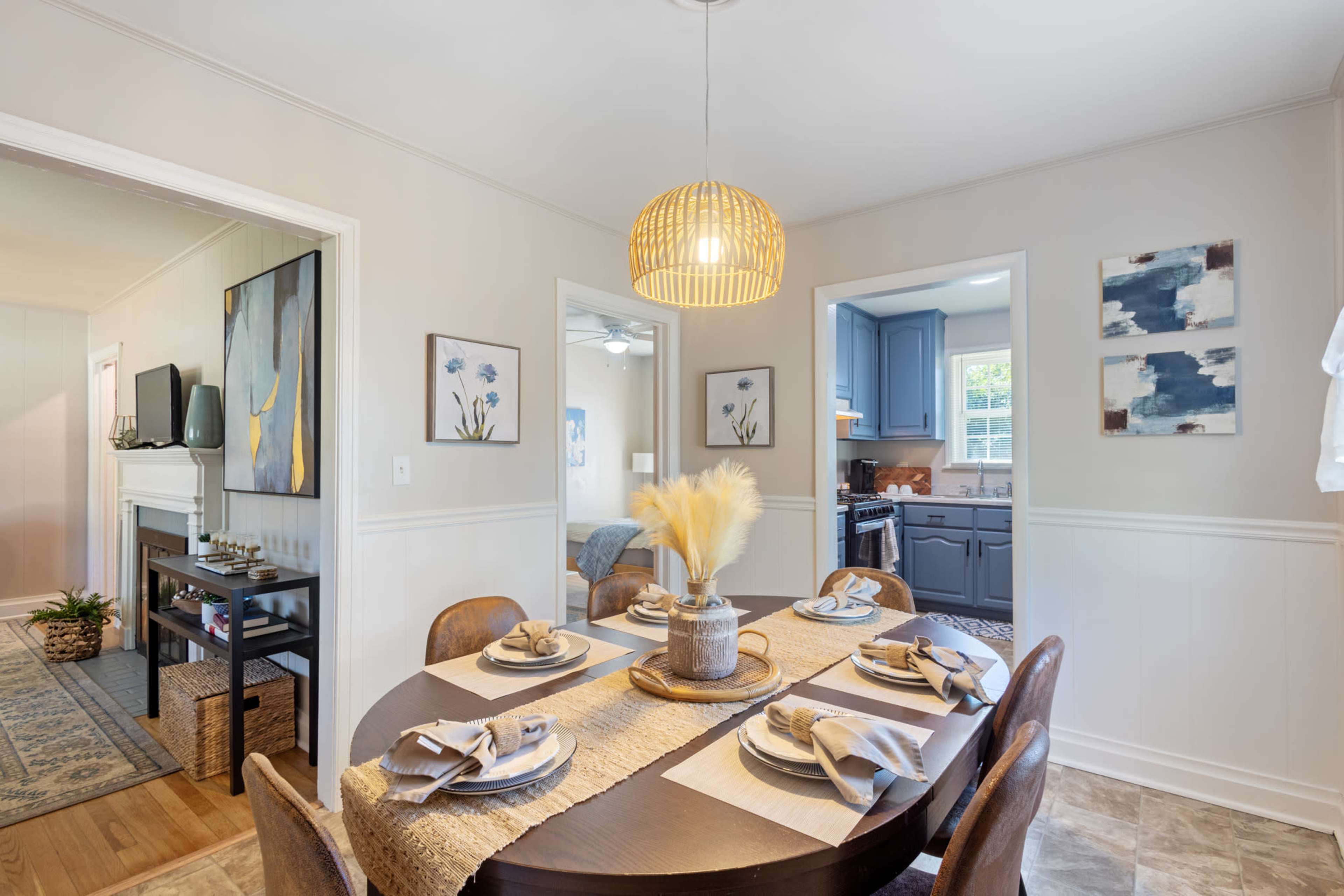 A dining area features a round table set with plates and utensils, surrounded by wooden chairs, with a view into a kitchen and living space.