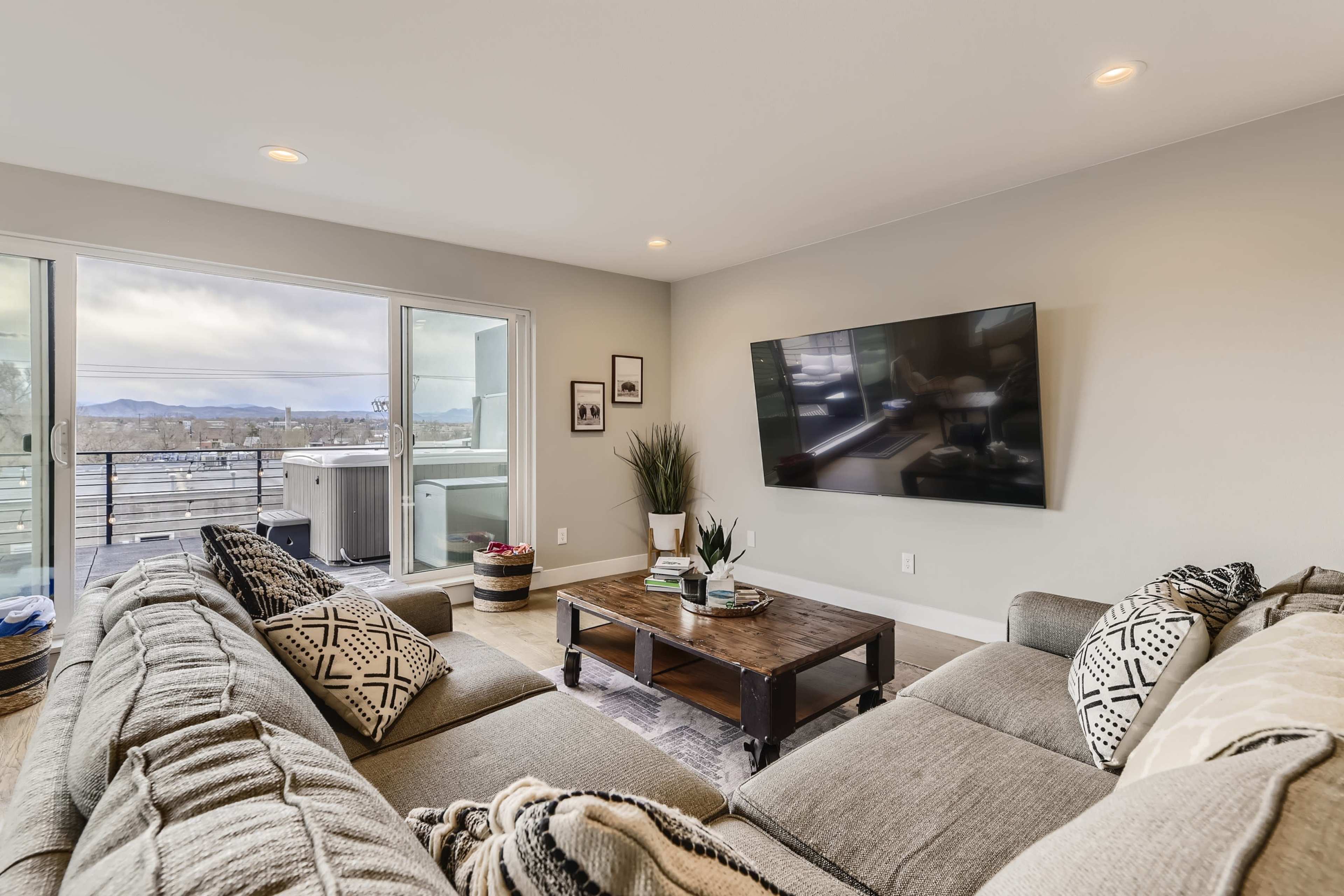 A modern living room features a large sectional sofa, a wooden coffee table, and a flat-screen TV mounted on the wall, with sliding glass doors leading to a balcony.