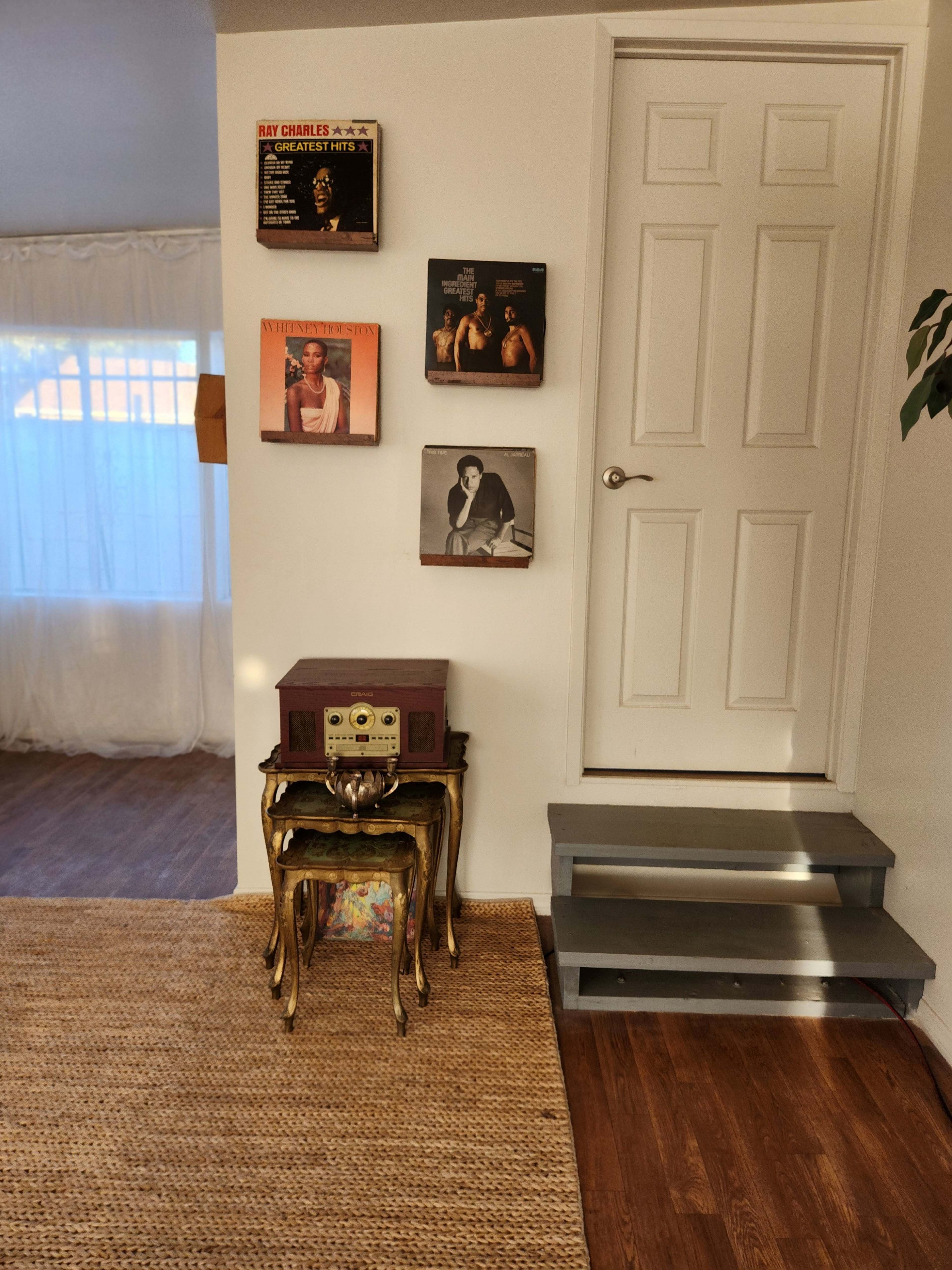 A small wooden table with a vintage record player sits next to a wall displaying several vinyl album covers, while a door leads to a staircase.