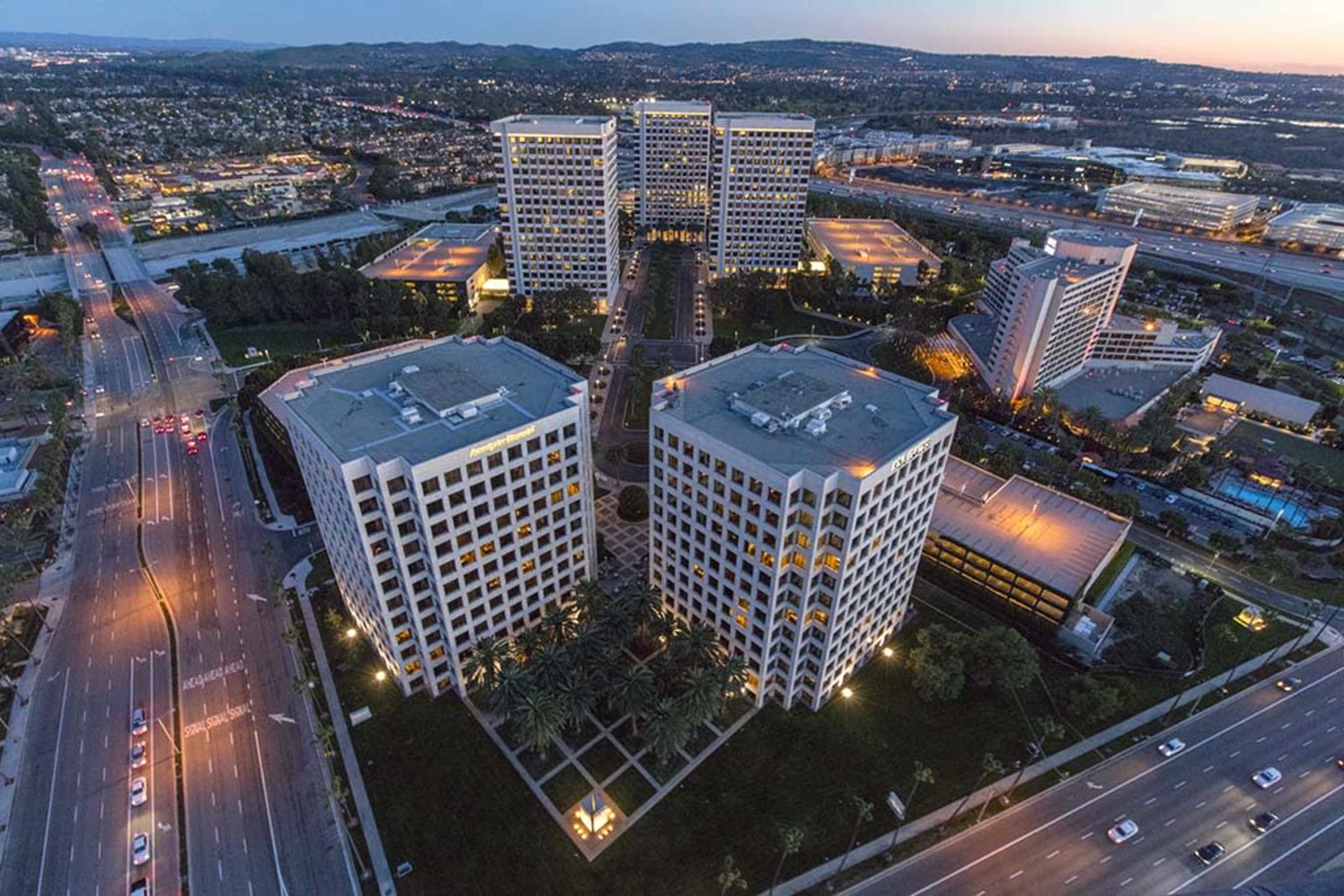 The image shows aerial view of several modern office buildings arranged in a landscaped area near a highway at dusk.