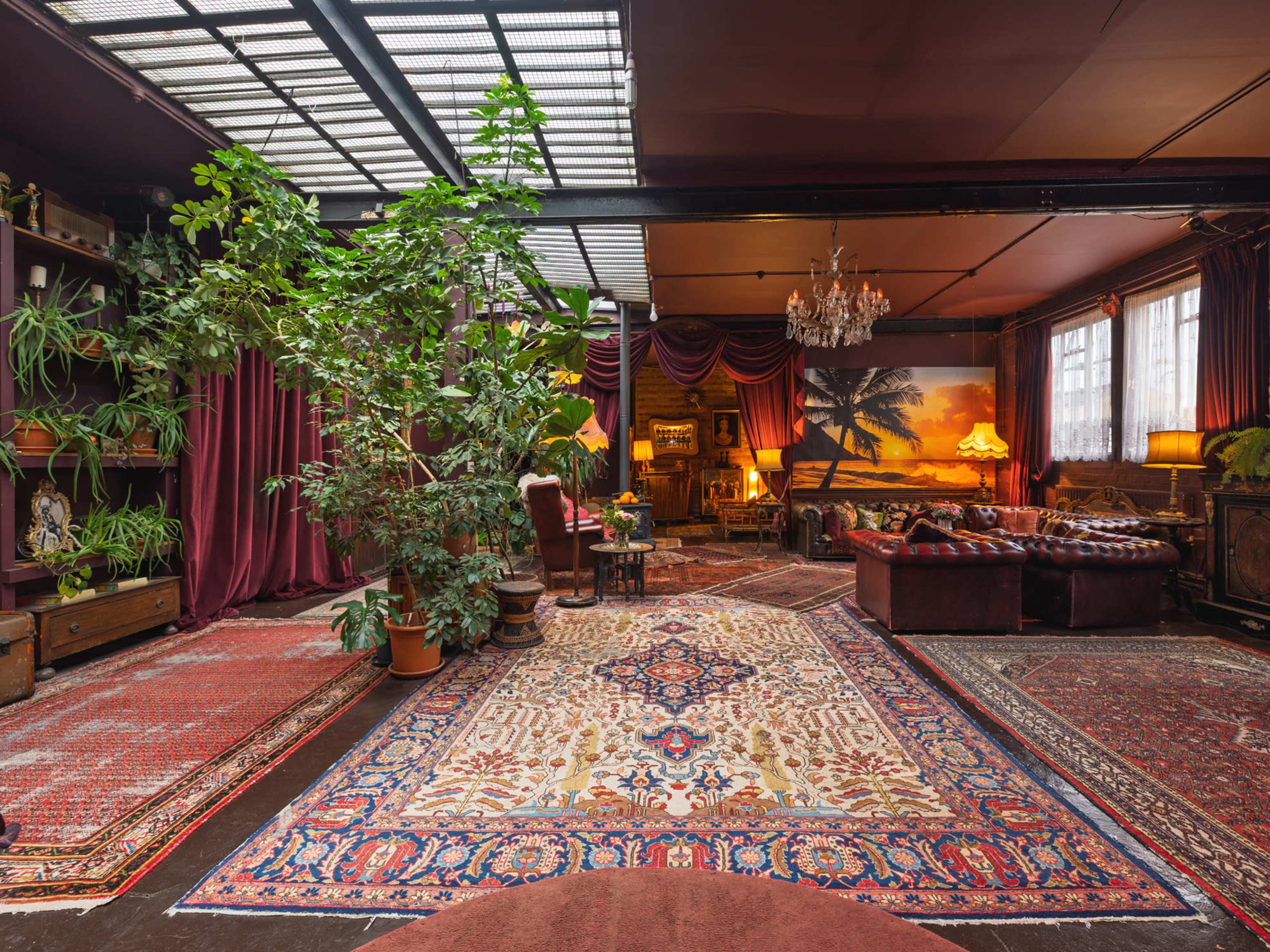 The image shows a spacious living area with rich red curtains, a large rug, potted plants, and vintage furniture under a glass roof.