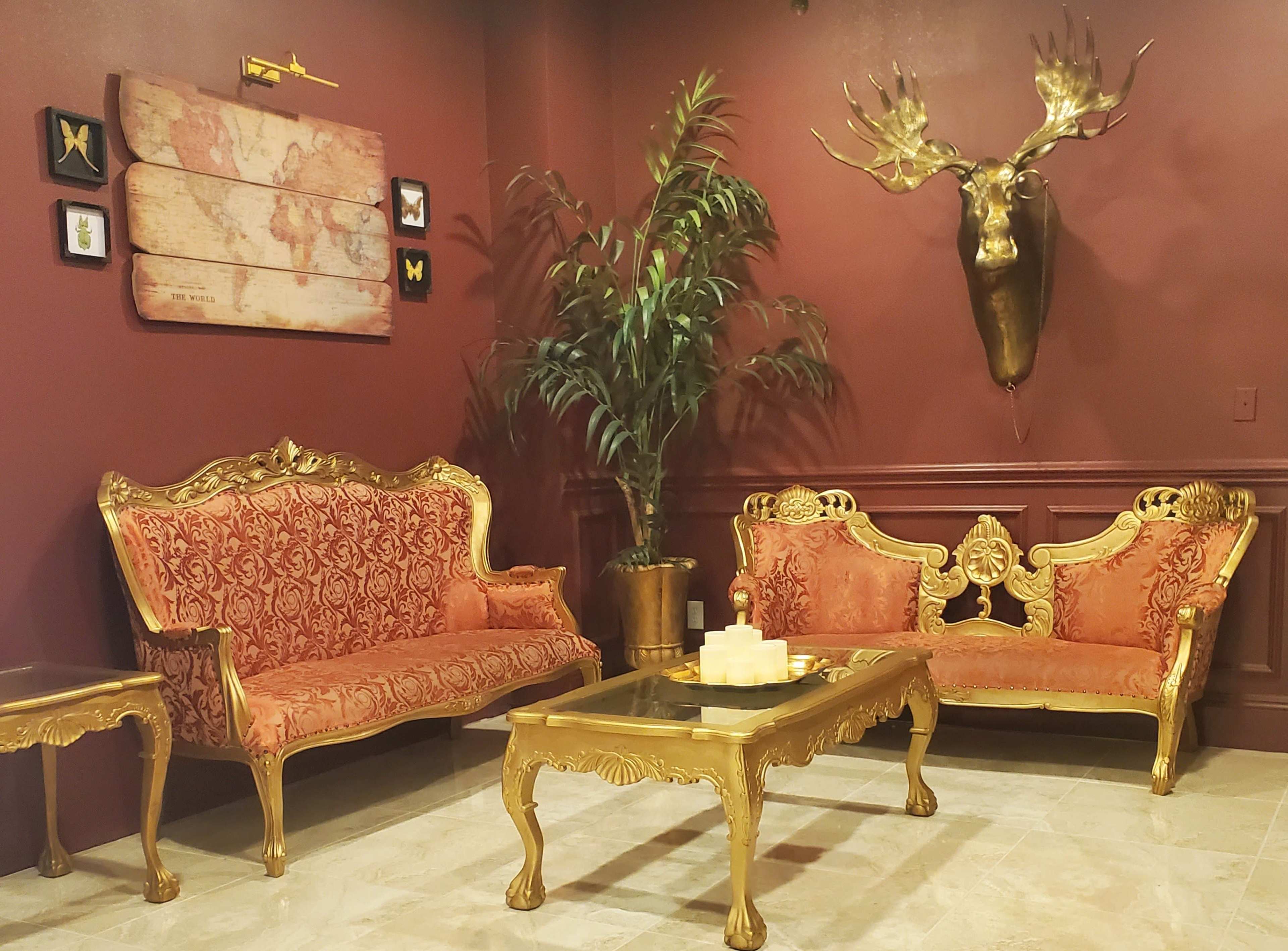 The image shows an ornate sitting area featuring gold furniture, plush orange upholstery, and a decorative wall with a world map and a moose head mount.