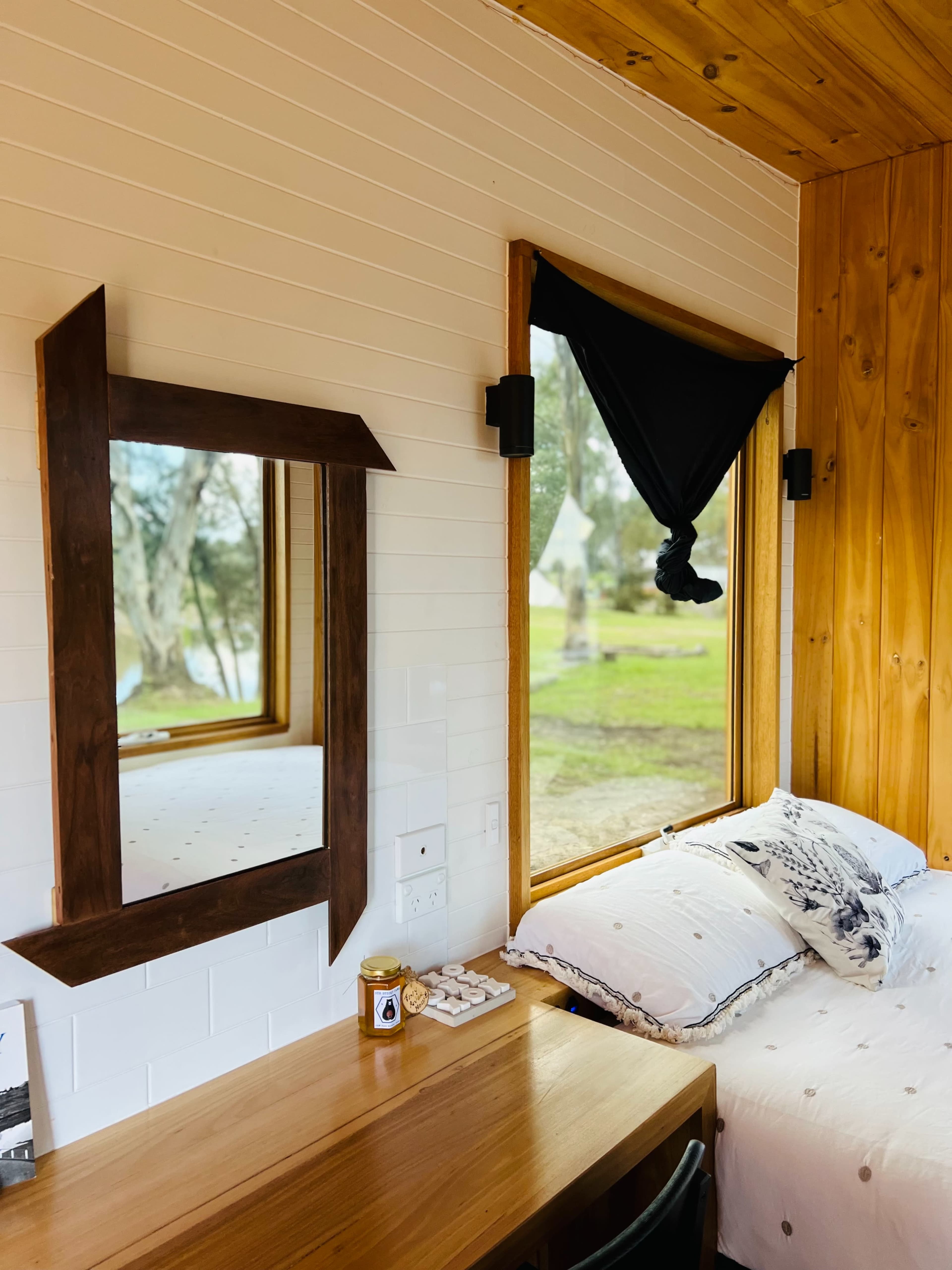 A wooden-framed mirror hangs on the wall beside a bed with white bedding, and a desk sits in front of a window with a black curtain.