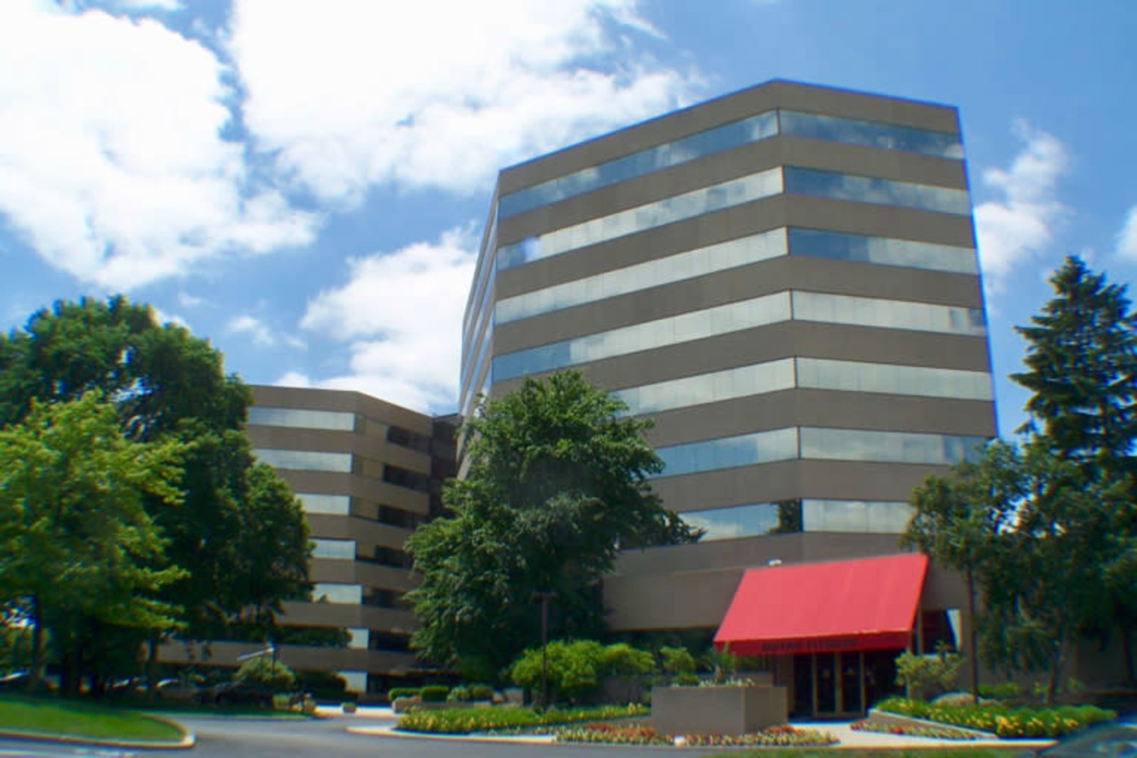 A modern office building with a distinctive red awning, surrounded by landscaped greenery under a partly cloudy sky.
