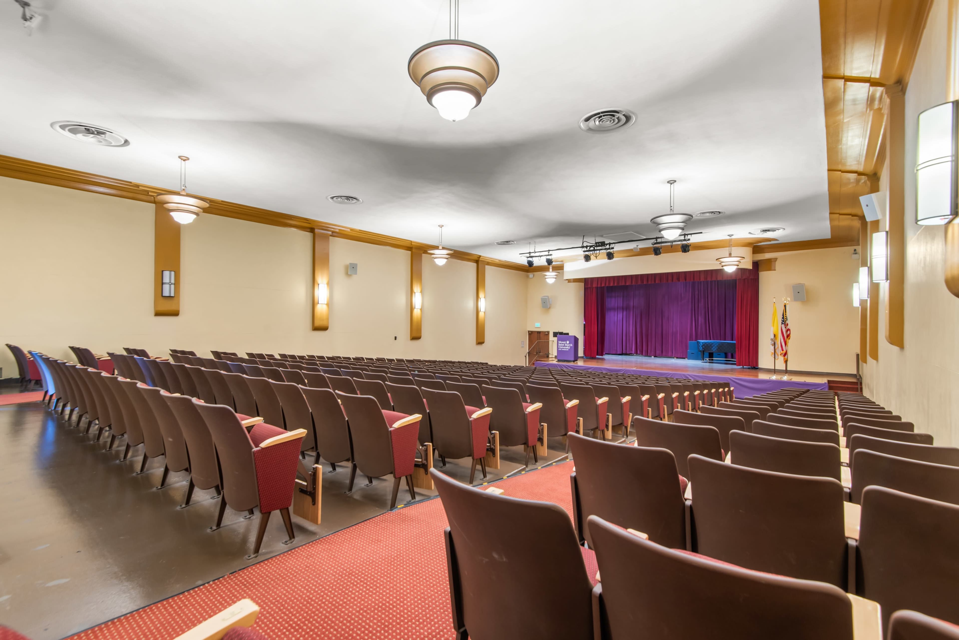 The image shows an empty auditorium with rows of brown chairs facing a purple stage, flanked by flags and equipped with stage lighting.