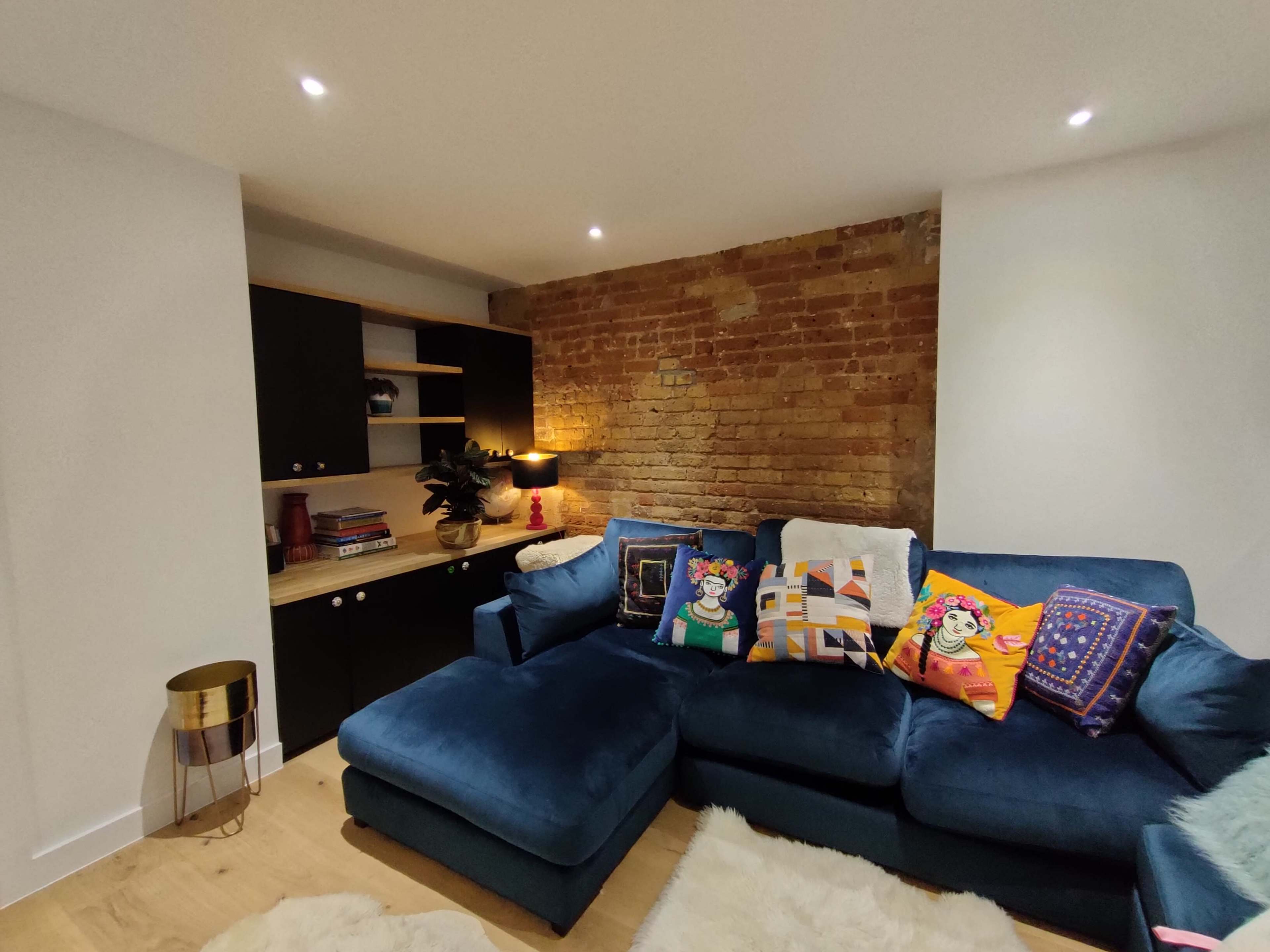 A blue sectional sofa with decorative pillows is positioned in a cozy living room featuring exposed brick walls and a beige carpet.