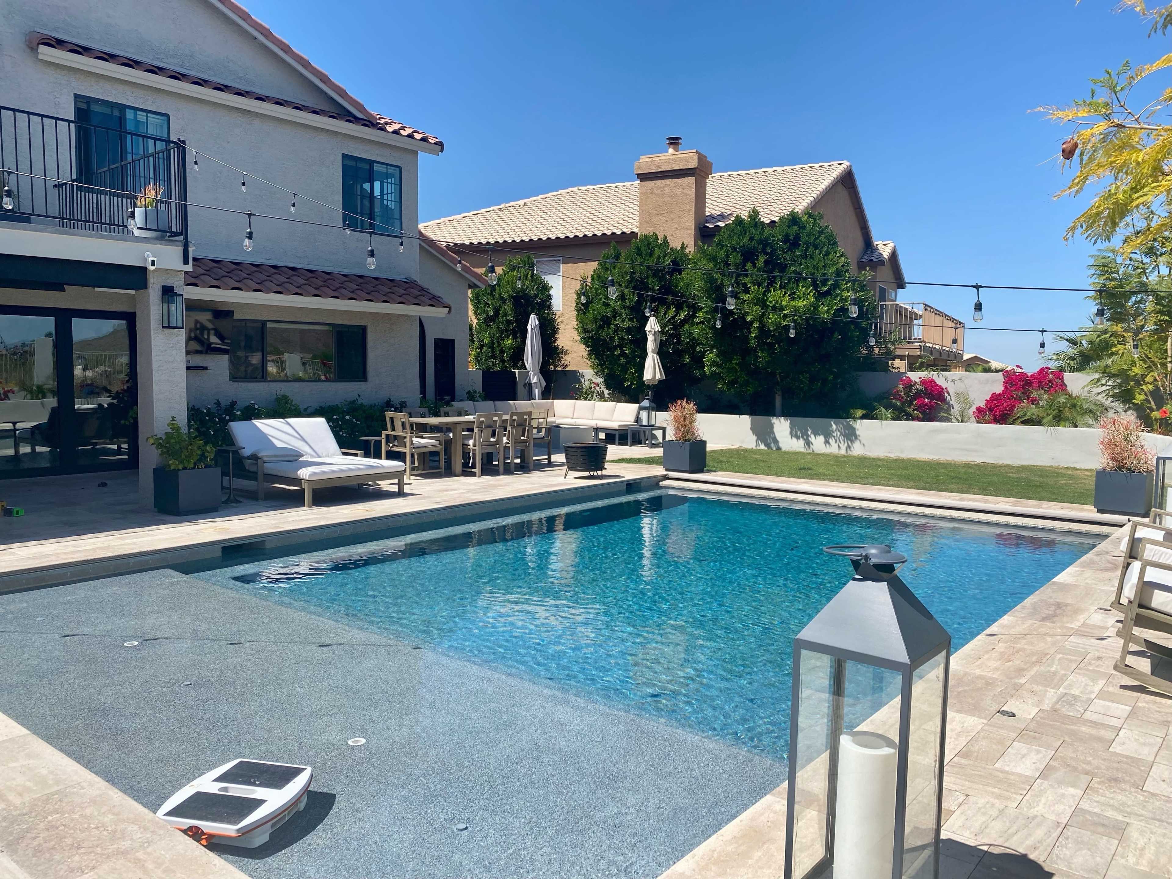 A backyard pool area featuring a clear swimming pool, lounge chairs, a dining table, and surrounding houses with landscaped vegetation.