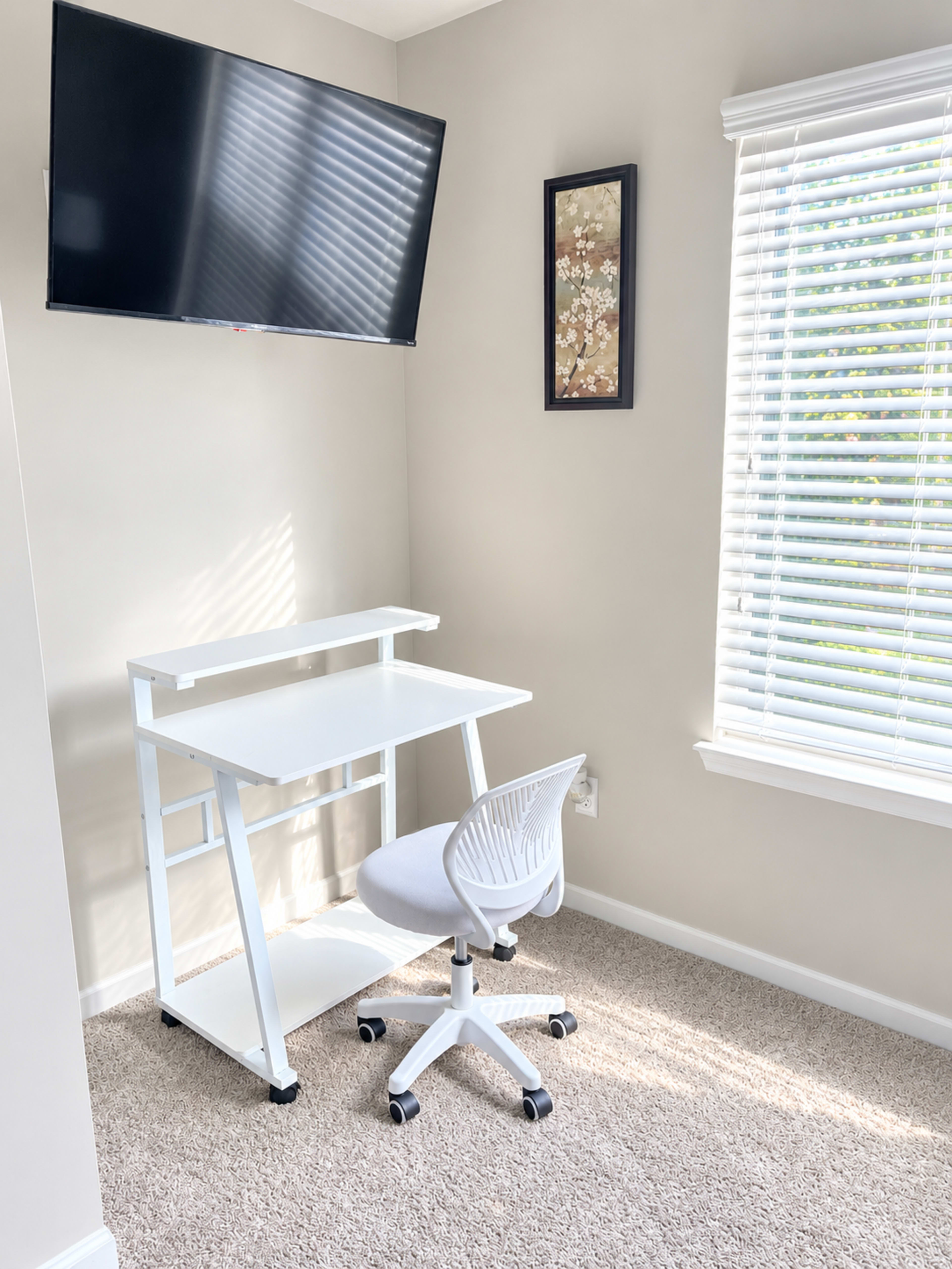 A minimalist workspace features a white desk and chair under a wall-mounted television, set against a neutral wall with a framed artwork and a window with blinds.