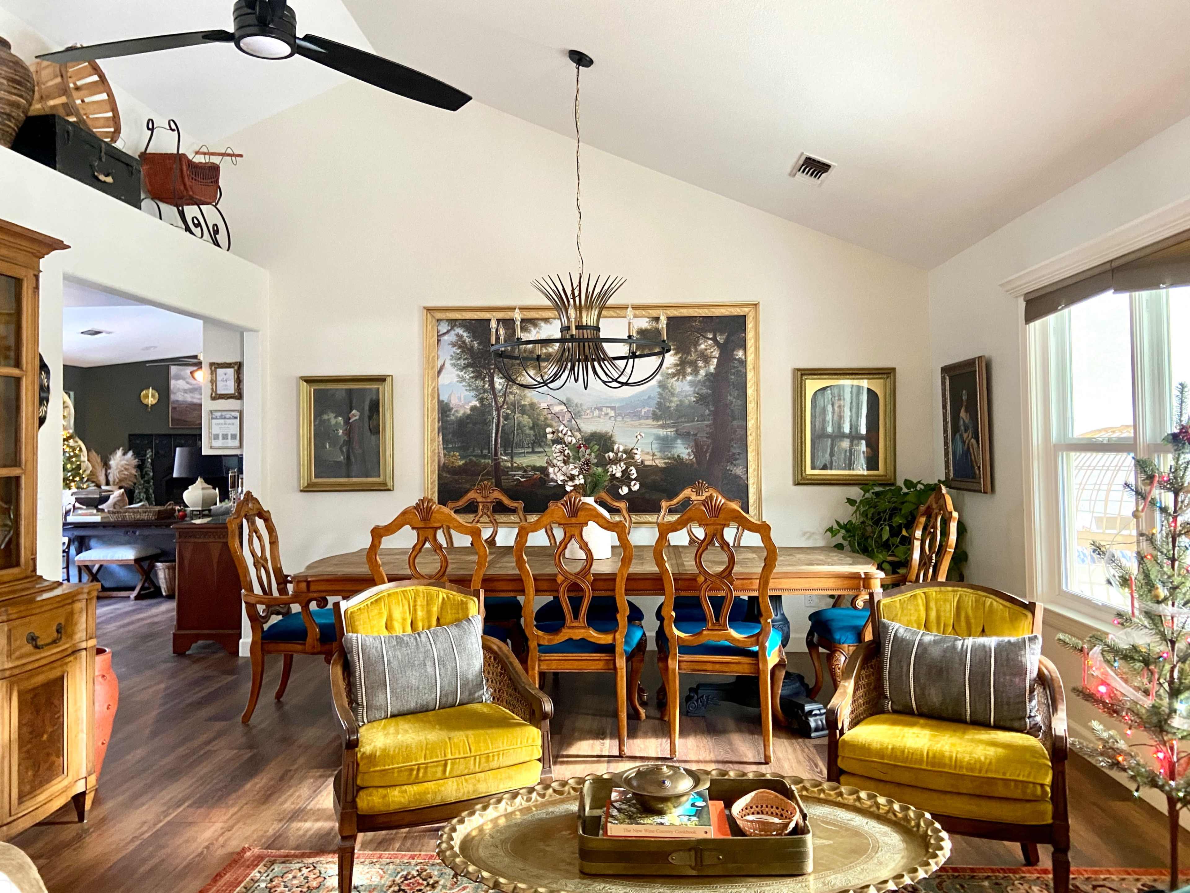 A dining area features a large wooden table surrounded by ornate chairs, accented by two yellow upholstered armchairs and a chandelier above.