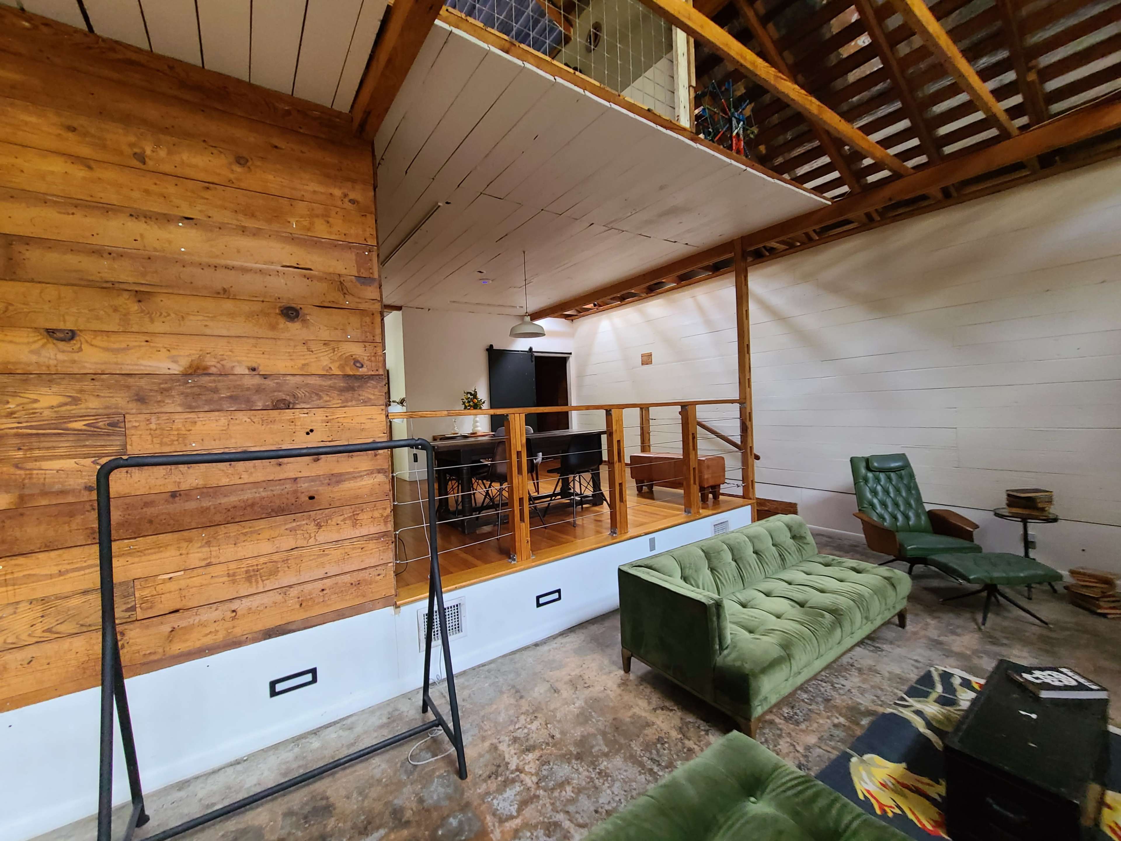 The image shows a living space with wooden walls, a green sofa, and a dining area featuring a table and chairs on an elevated platform.