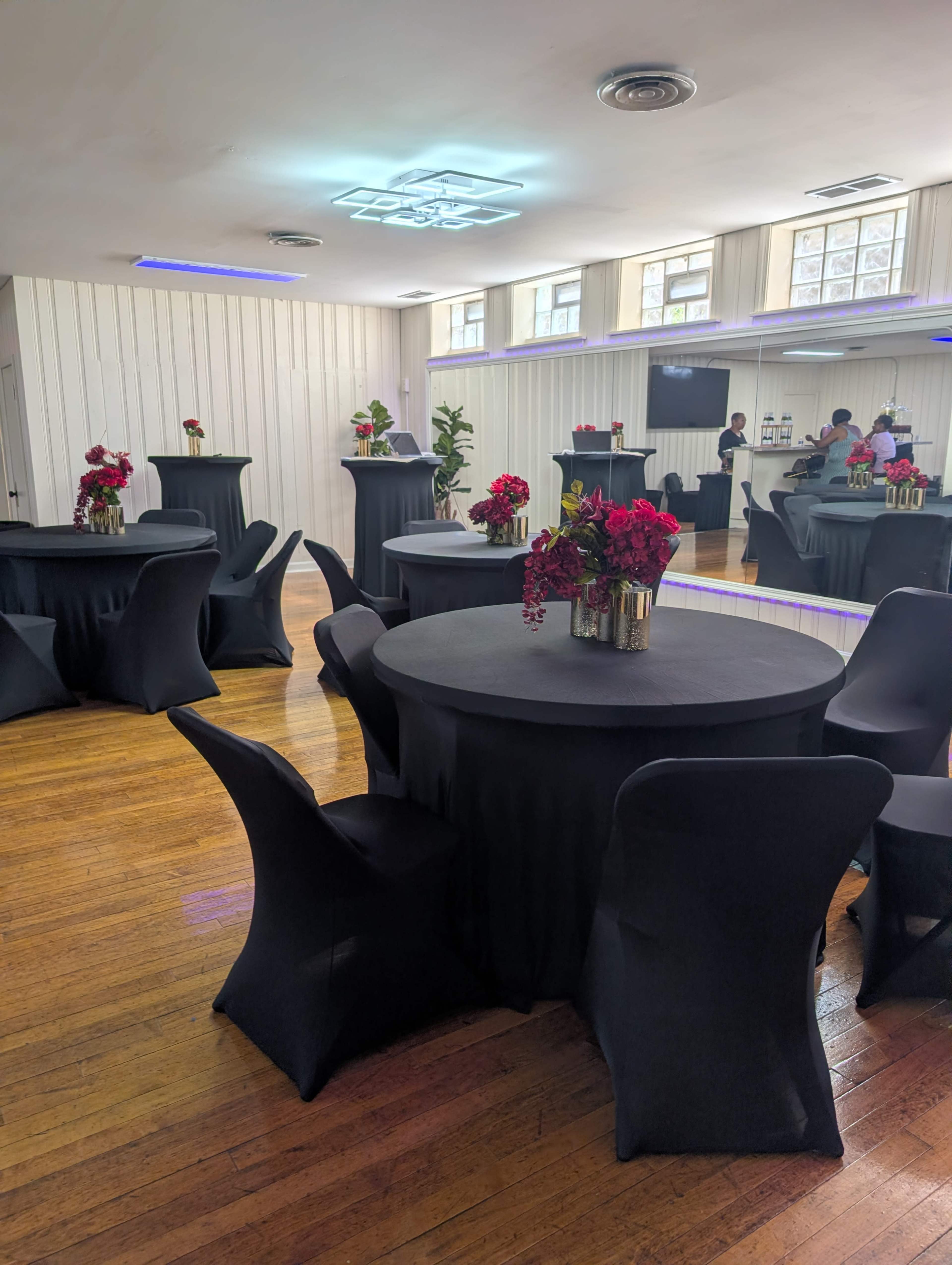 The image shows a spacious event room with black tablecloth-covered round tables adorned with floral centerpieces and sleek black chairs.