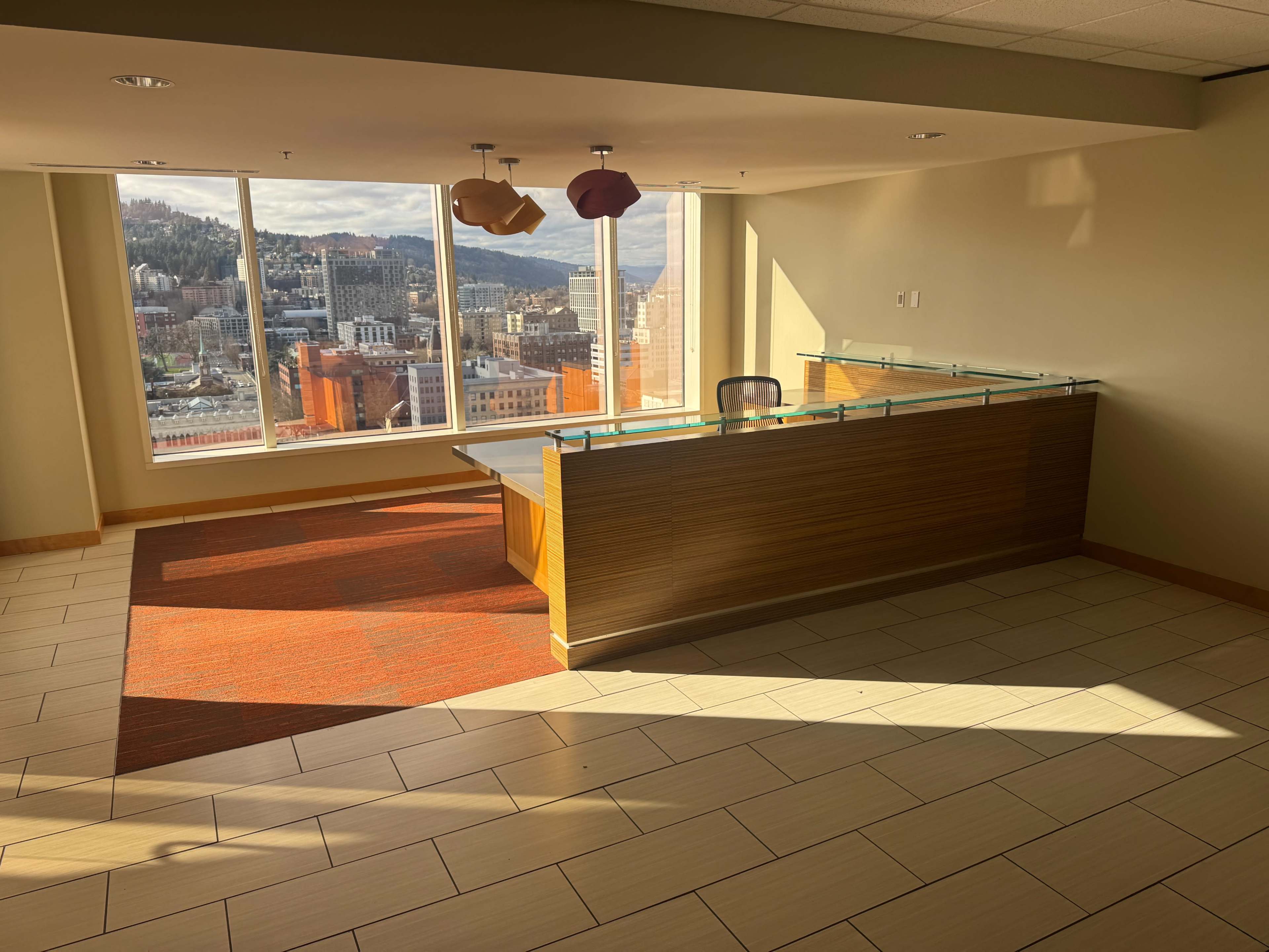 The image shows a modern reception area with a wooden front desk, large windows offering a view of a cityscape, and colorful light fixtures hanging from the ceiling.