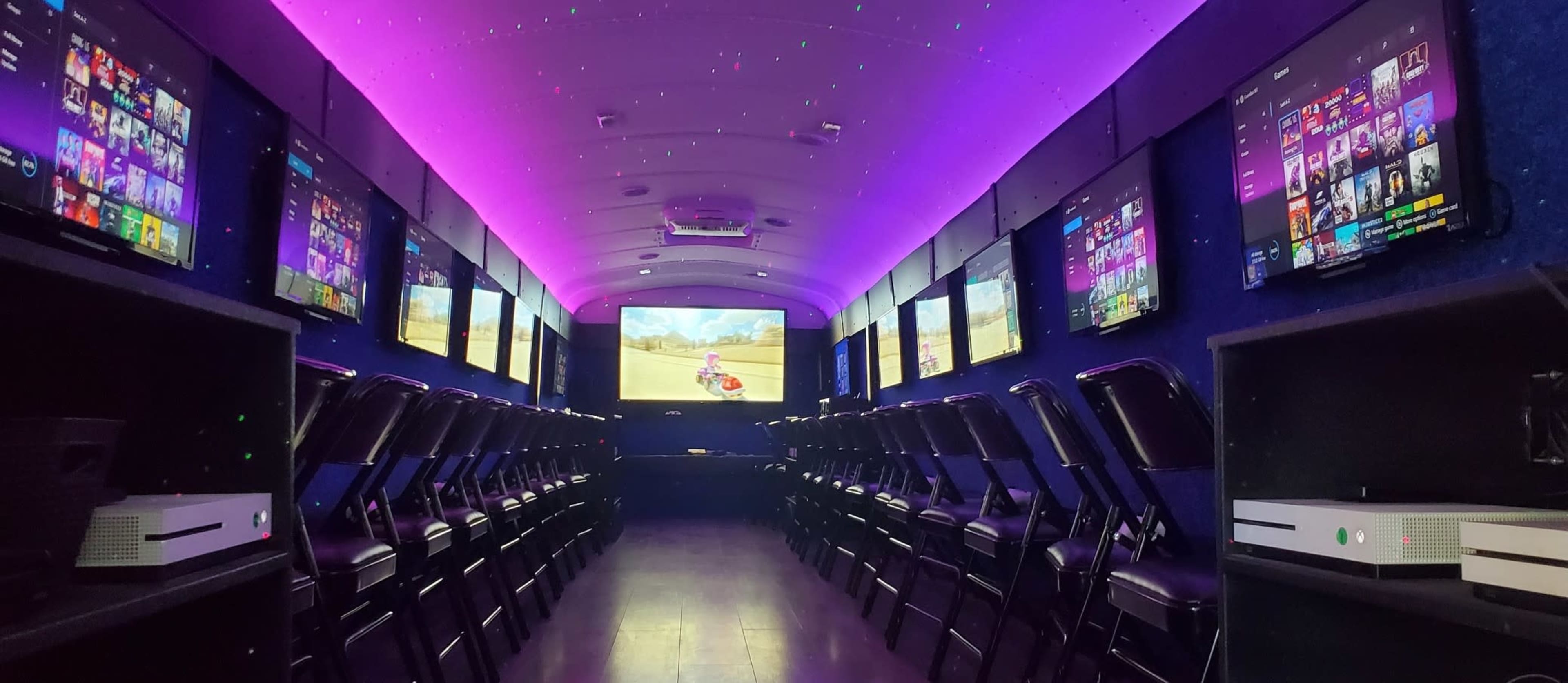 The image shows an interior of a gaming room or bus equipped with multiple screens and chairs arranged in rows under purple ambient lighting.