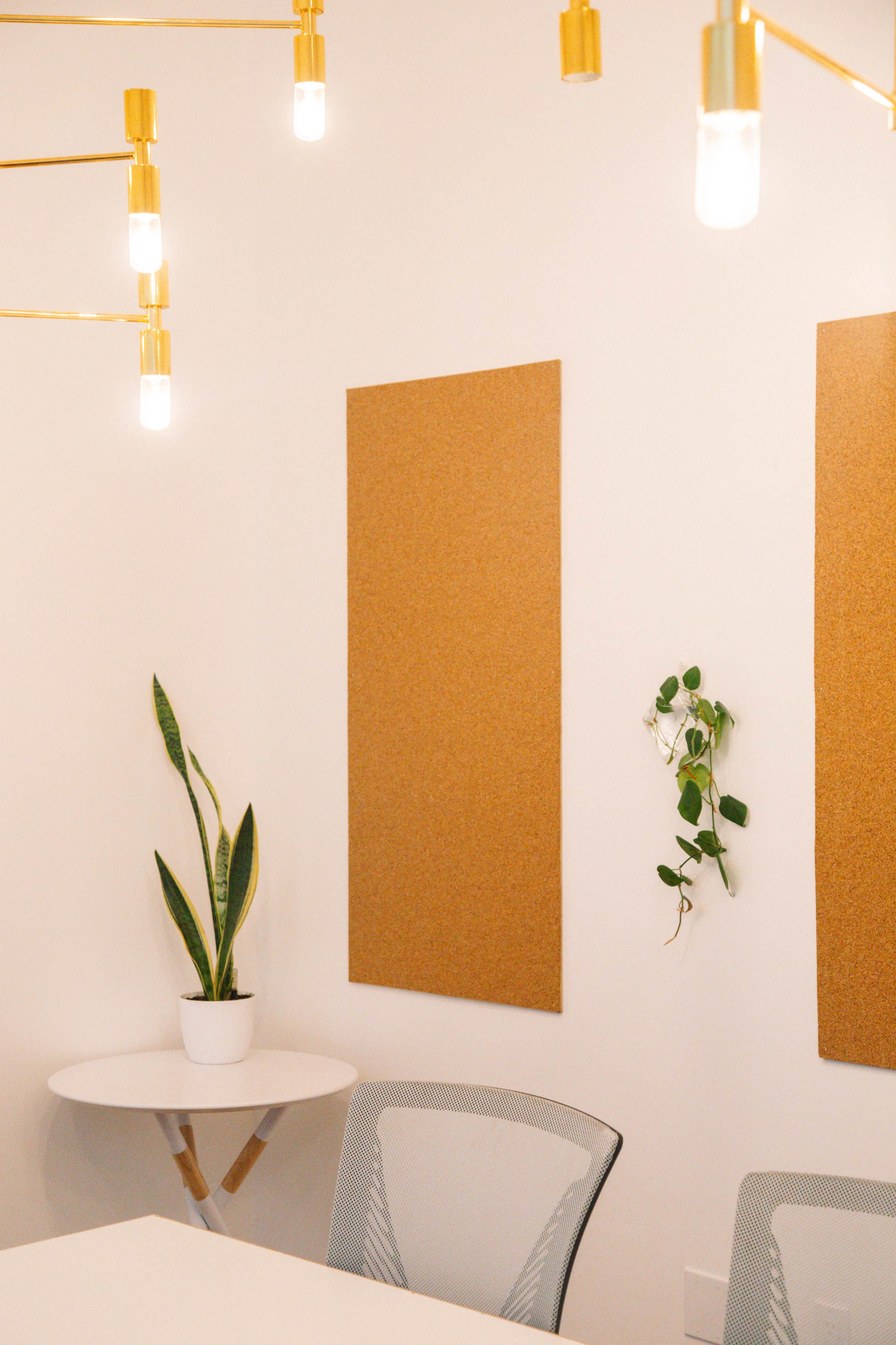 The image shows a minimalistic meeting room with a round white table, a plant in a pot, cork boards on the walls, and pendant lights hanging from the ceiling.