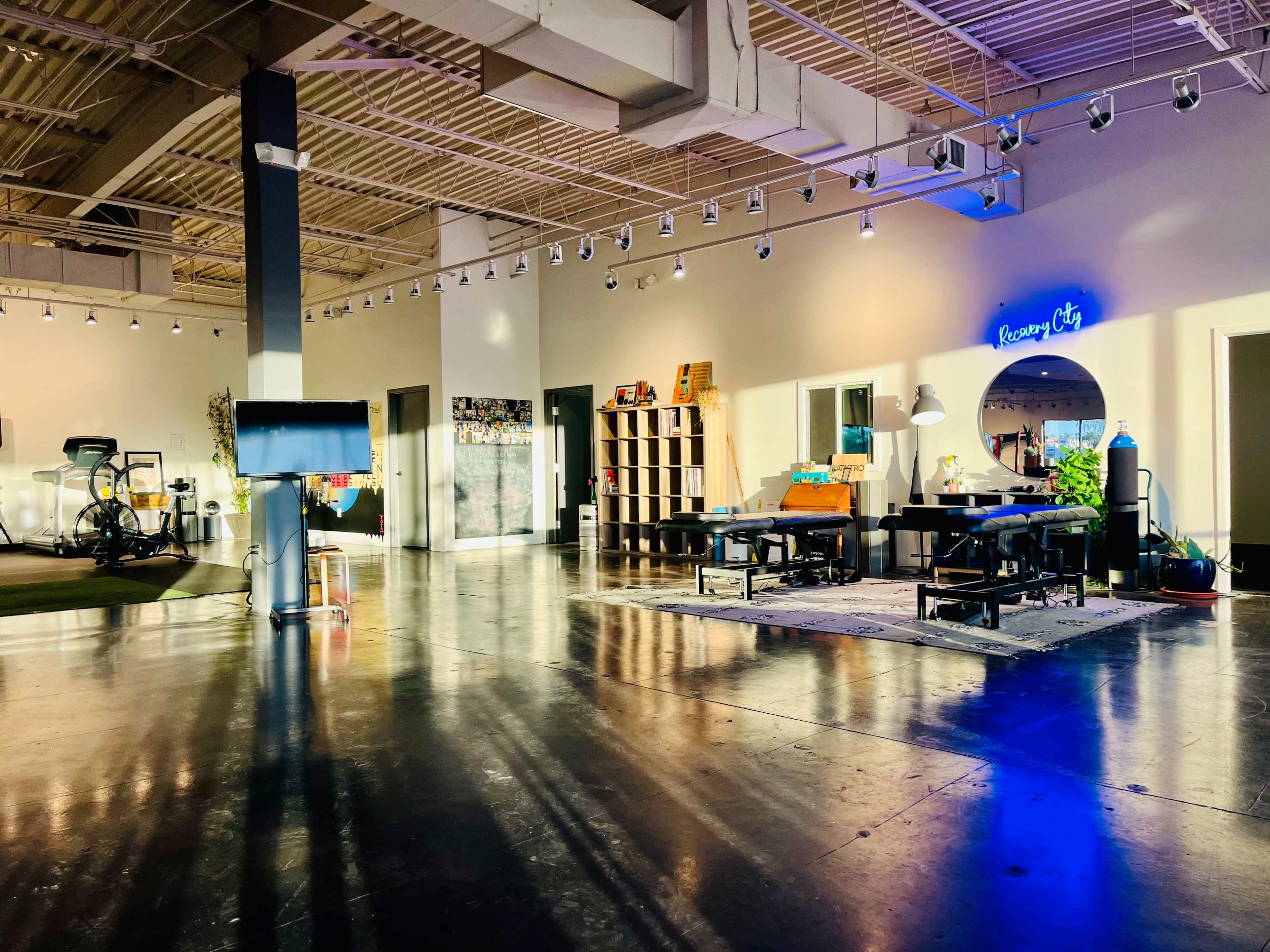 An open, well-lit space featuring workout equipment, seating areas, and decorative elements, with large windows casting shadows on the polished floor.