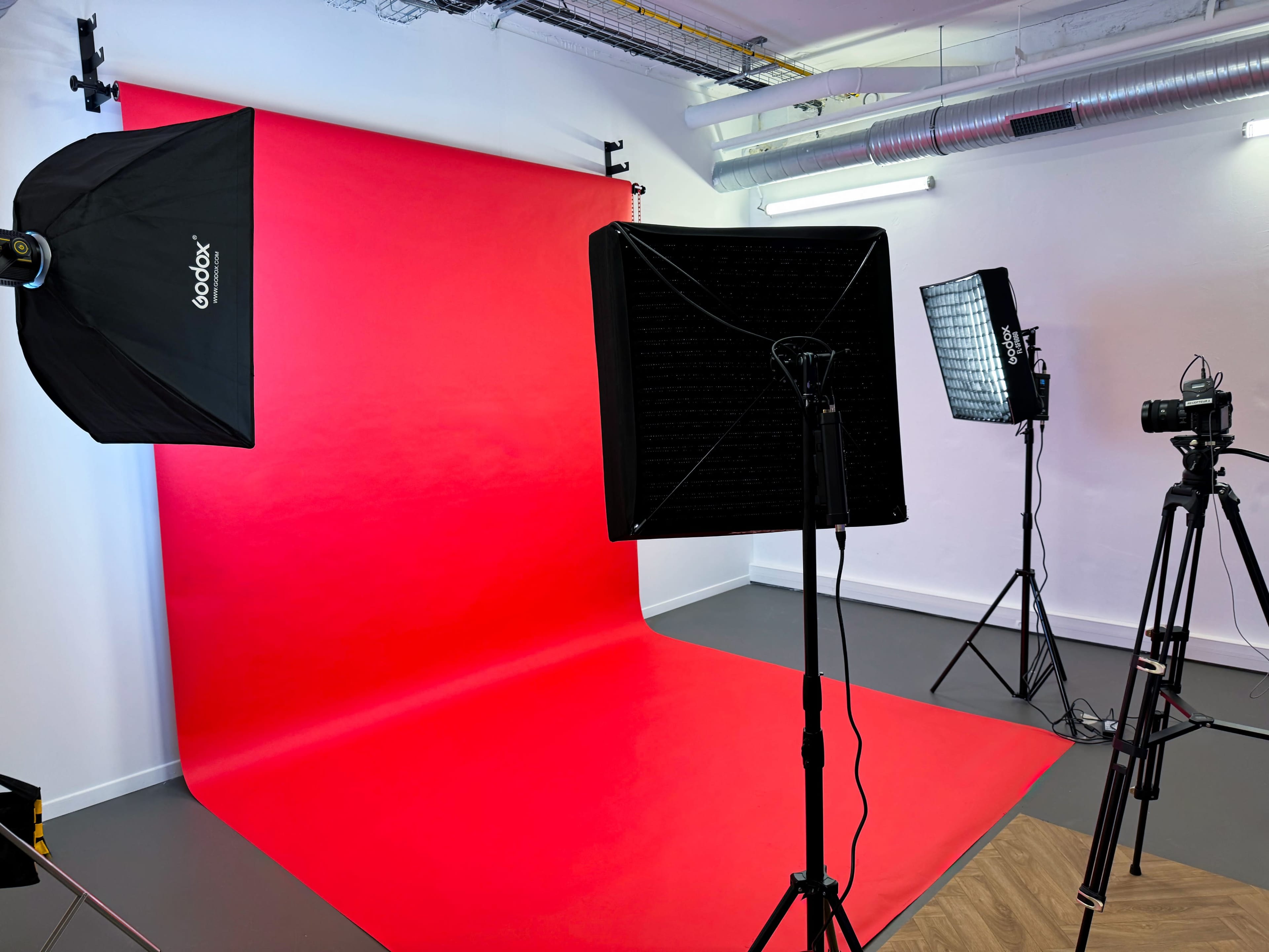 The image shows a photography studio with a red backdrop, two light sources, and a camera setup.