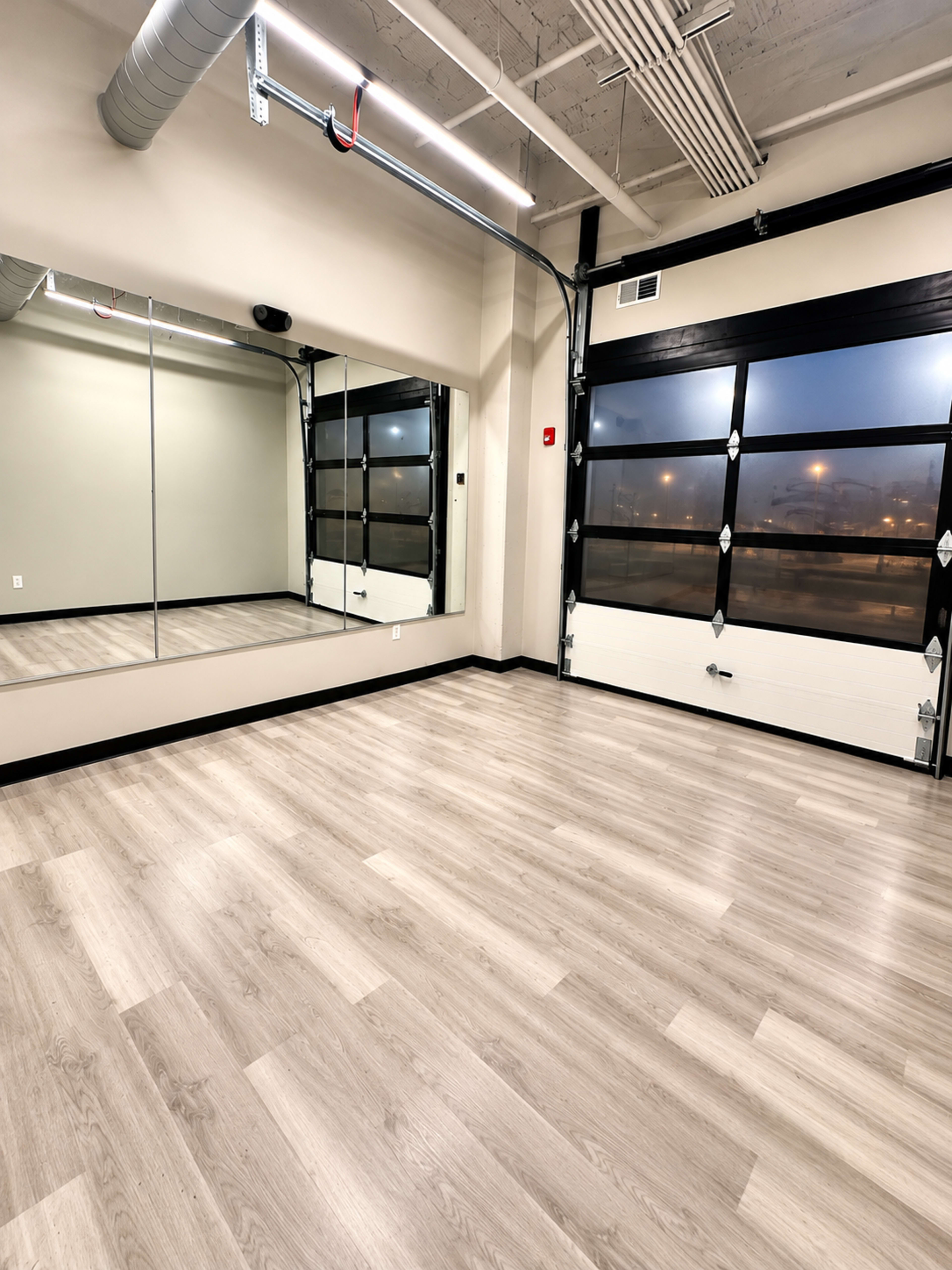 The image shows a spacious, empty room with wooden flooring, large mirrors on one wall, and a garage door window letting in ambient light.