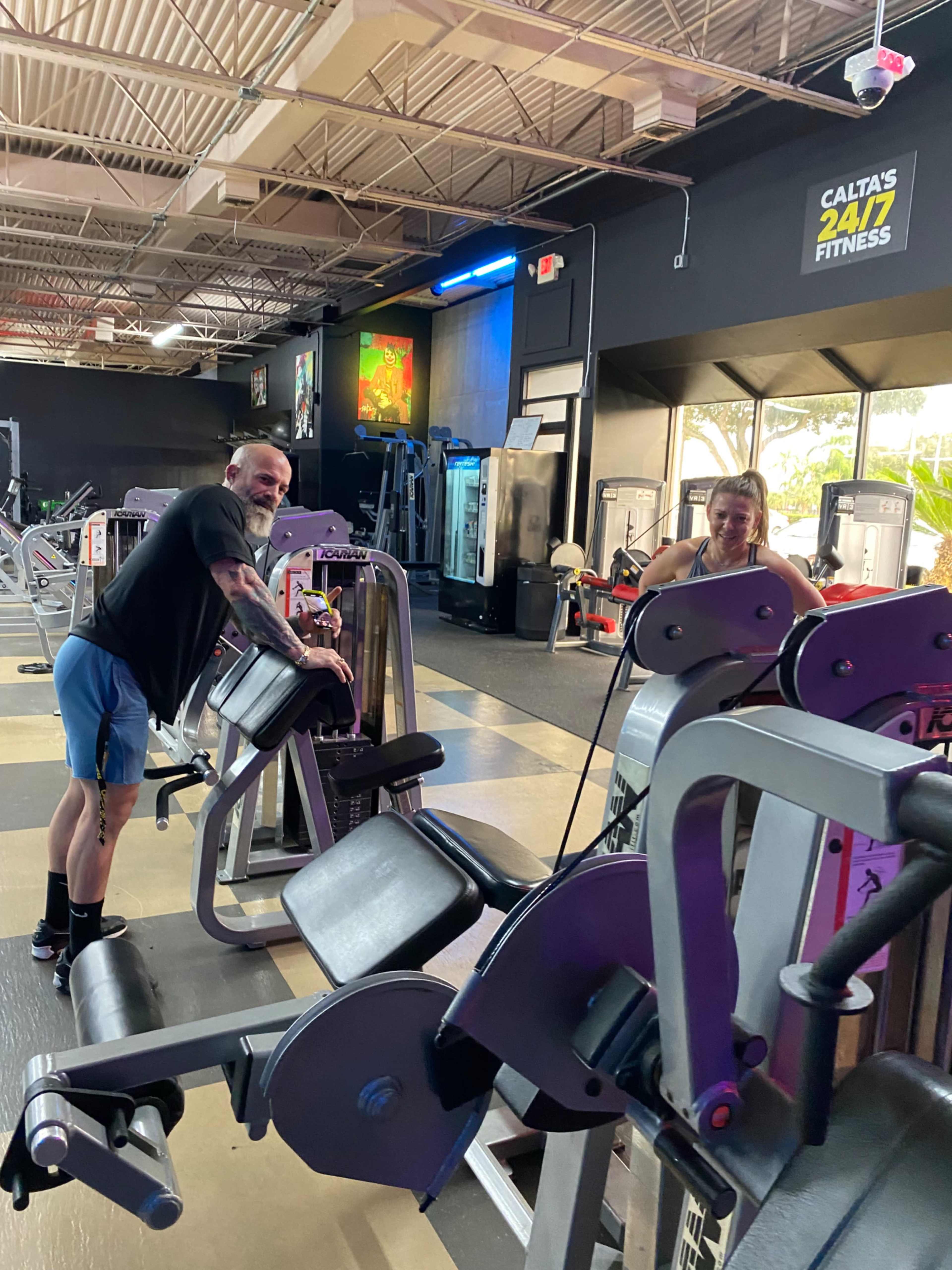 Two people are using gym equipment in a fitness center with various machines in the background.