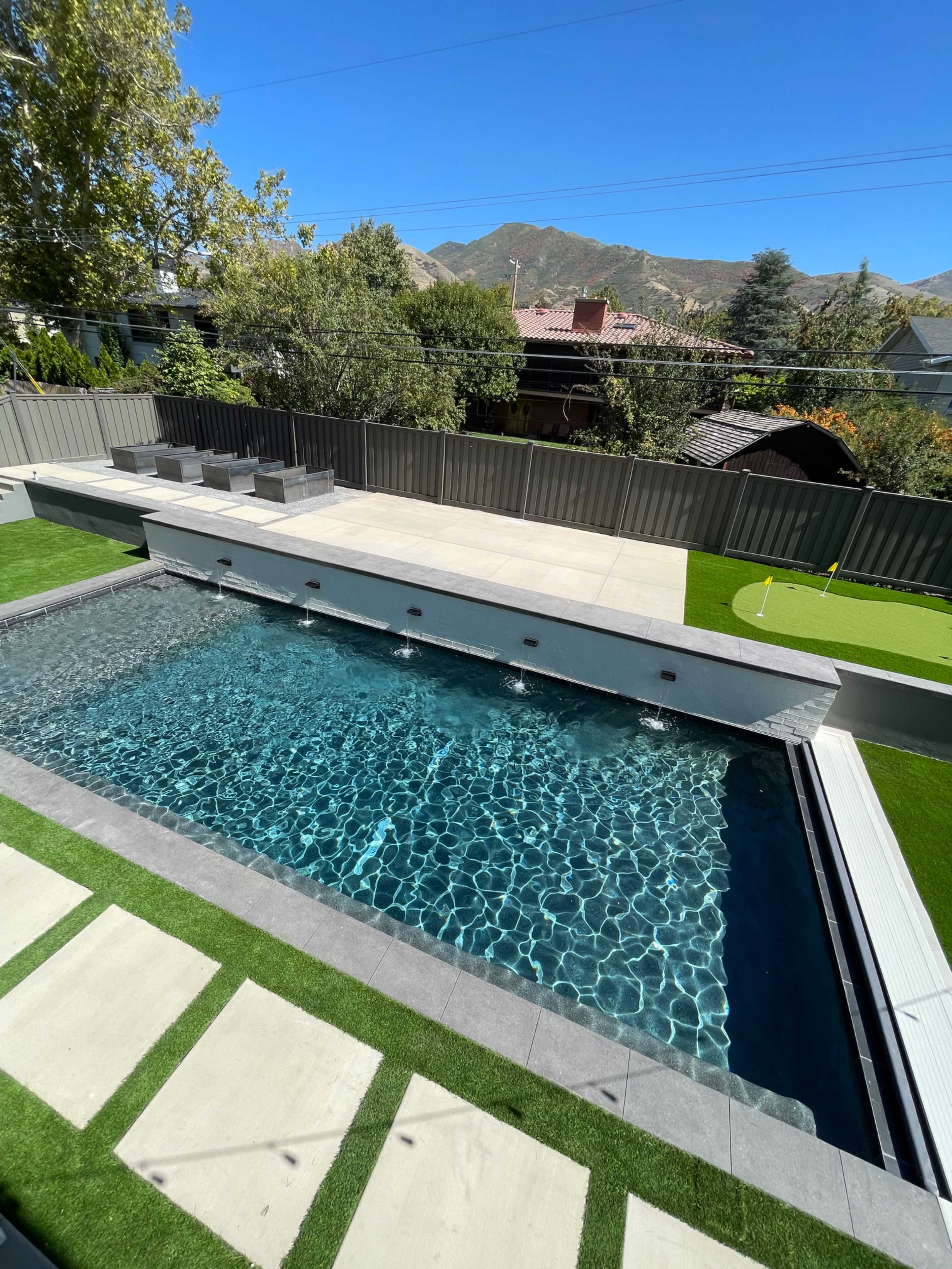 A modern backyard featuring a rectangular swimming pool with clear water, surrounded by concrete decking and artificial grass, with mountains in the background.