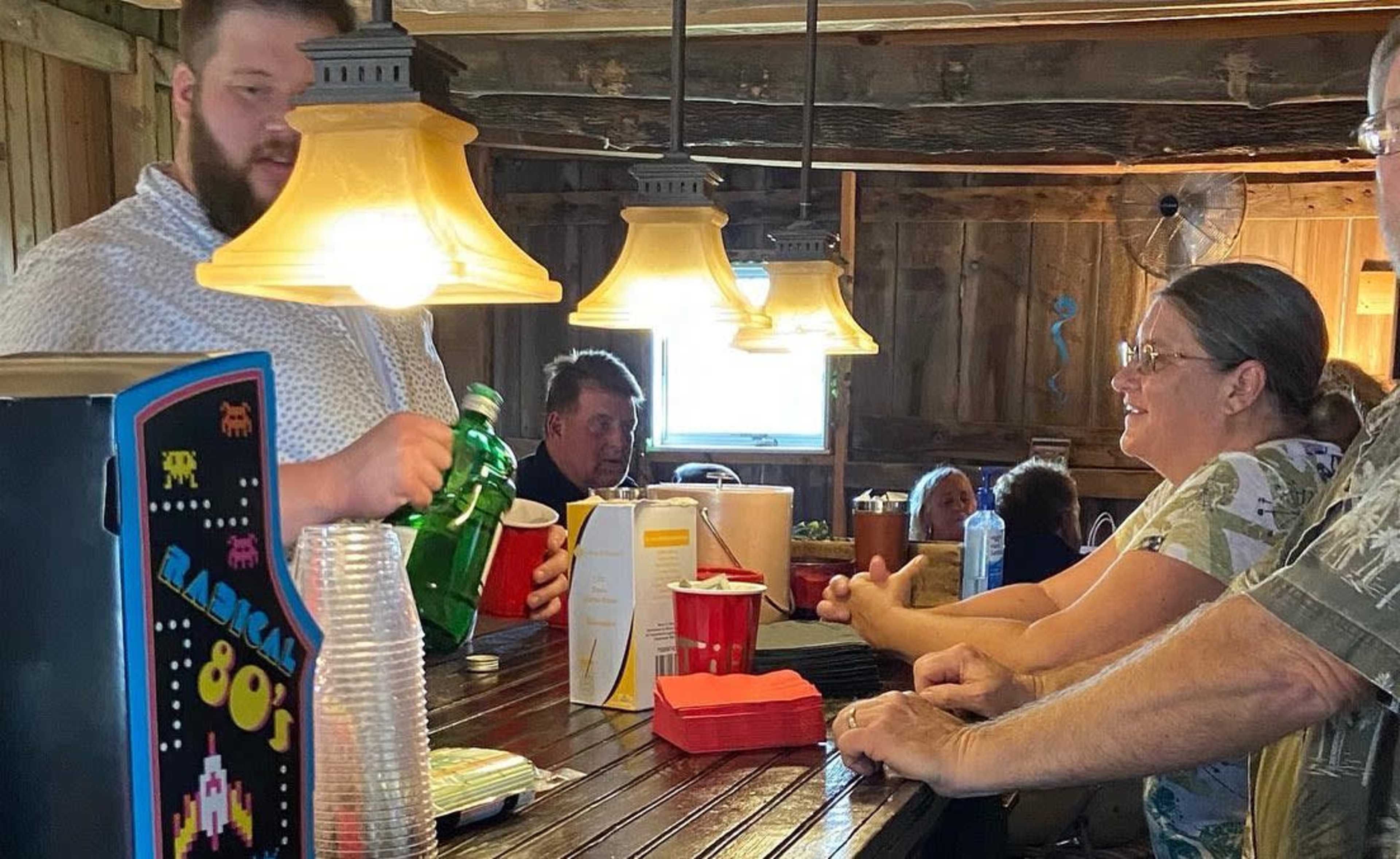 A bartender pours a drink for a patron at a rustic wooden bar, while several customers engage in conversation nearby.