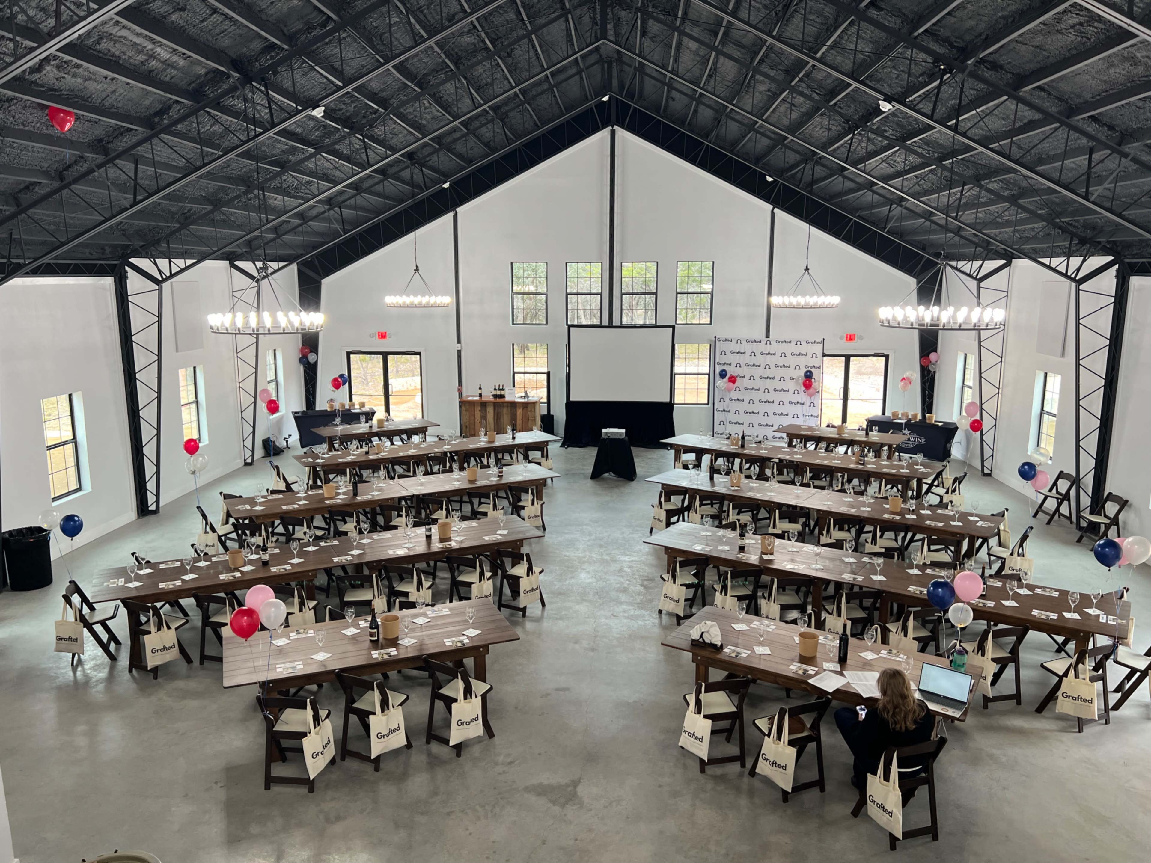 The image shows a spacious indoor event venue with multiple rows of tables set for a gathering, adorned with balloons and a presentation screen at the front.
