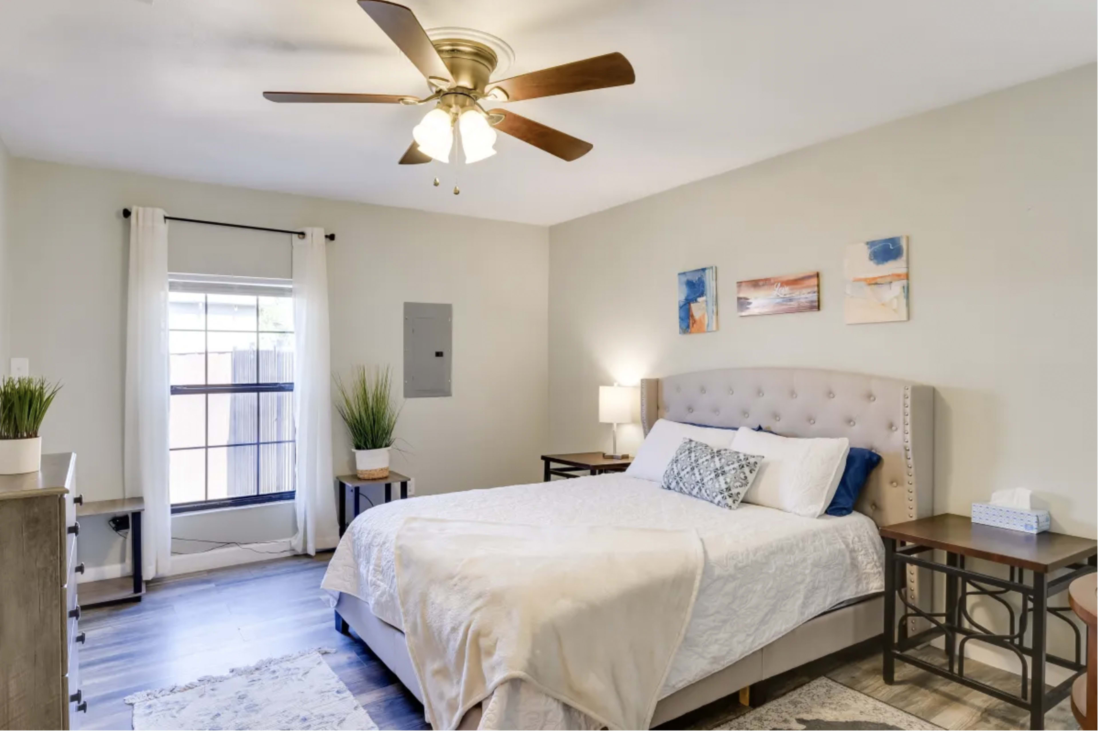 A neatly arranged bedroom featuring a queen-sized bed with a tufted headboard, a pair of bedside tables, and a window with sheer curtains.