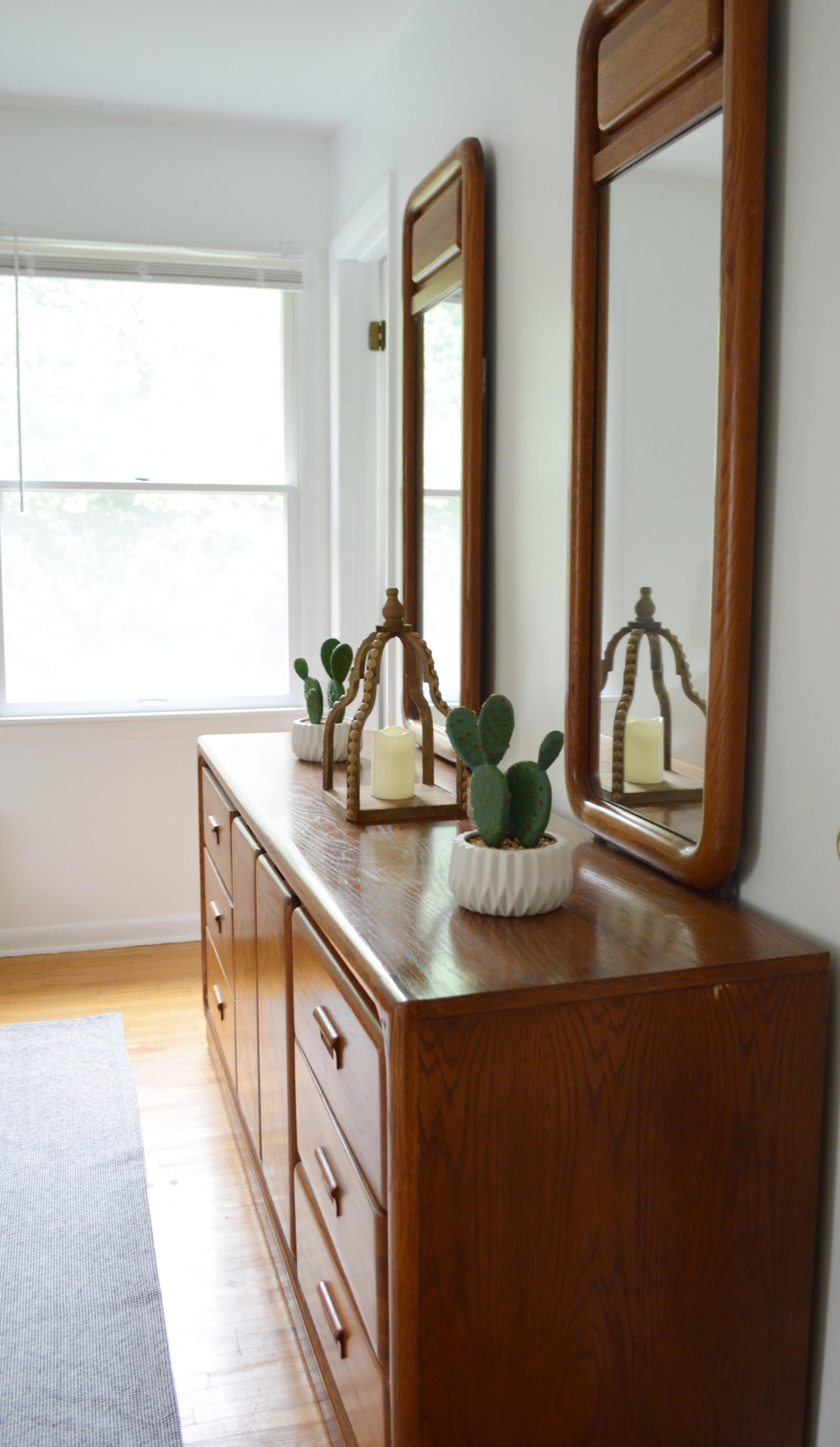 A wooden dresser with drawers and two mirrors is positioned against a wall, adorned with a lantern and potted cacti.