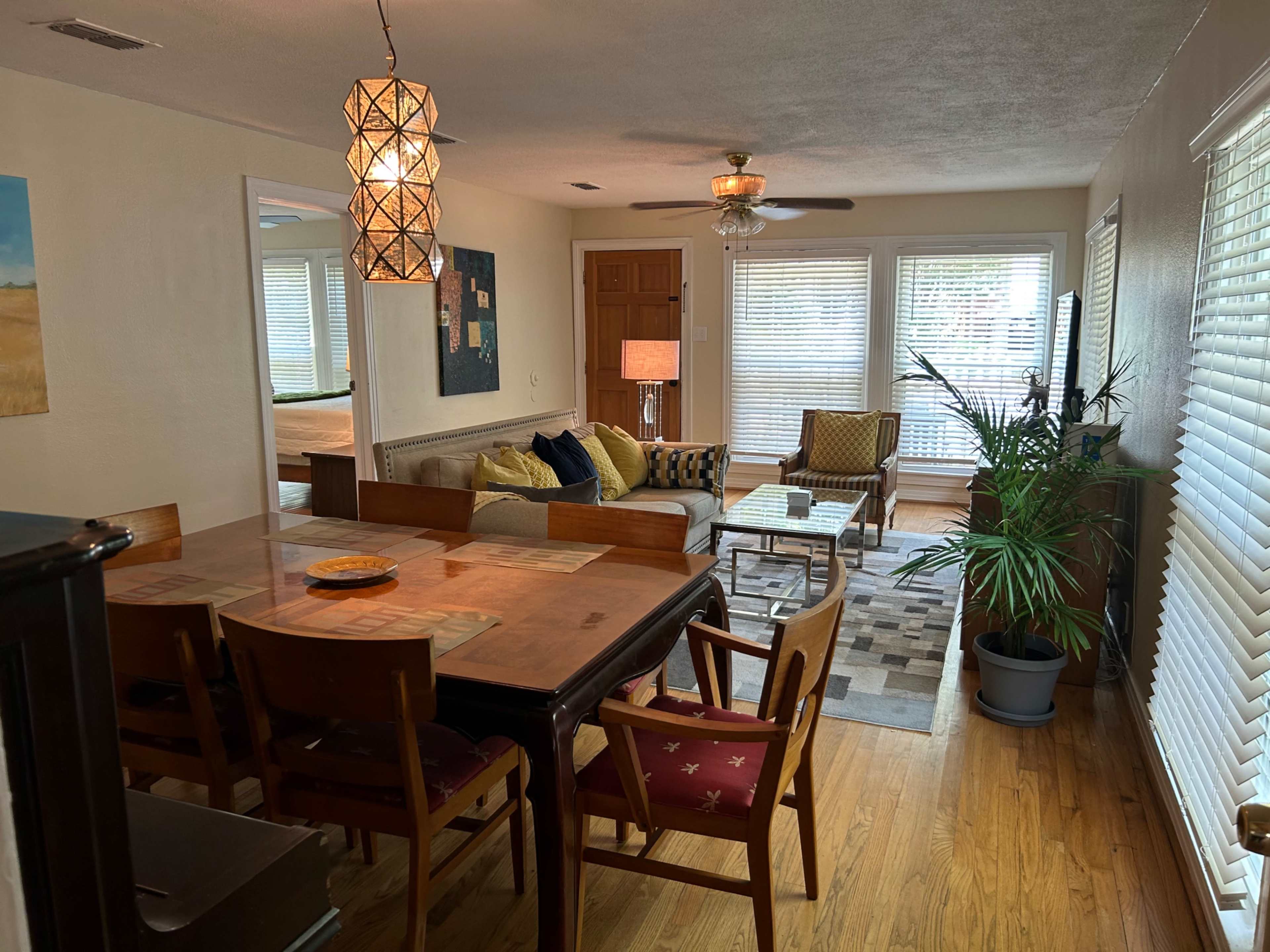 The image shows a bright and inviting living area with a dining table, comfortable seating in the lounge, and several windows allowing natural light to fill the space.