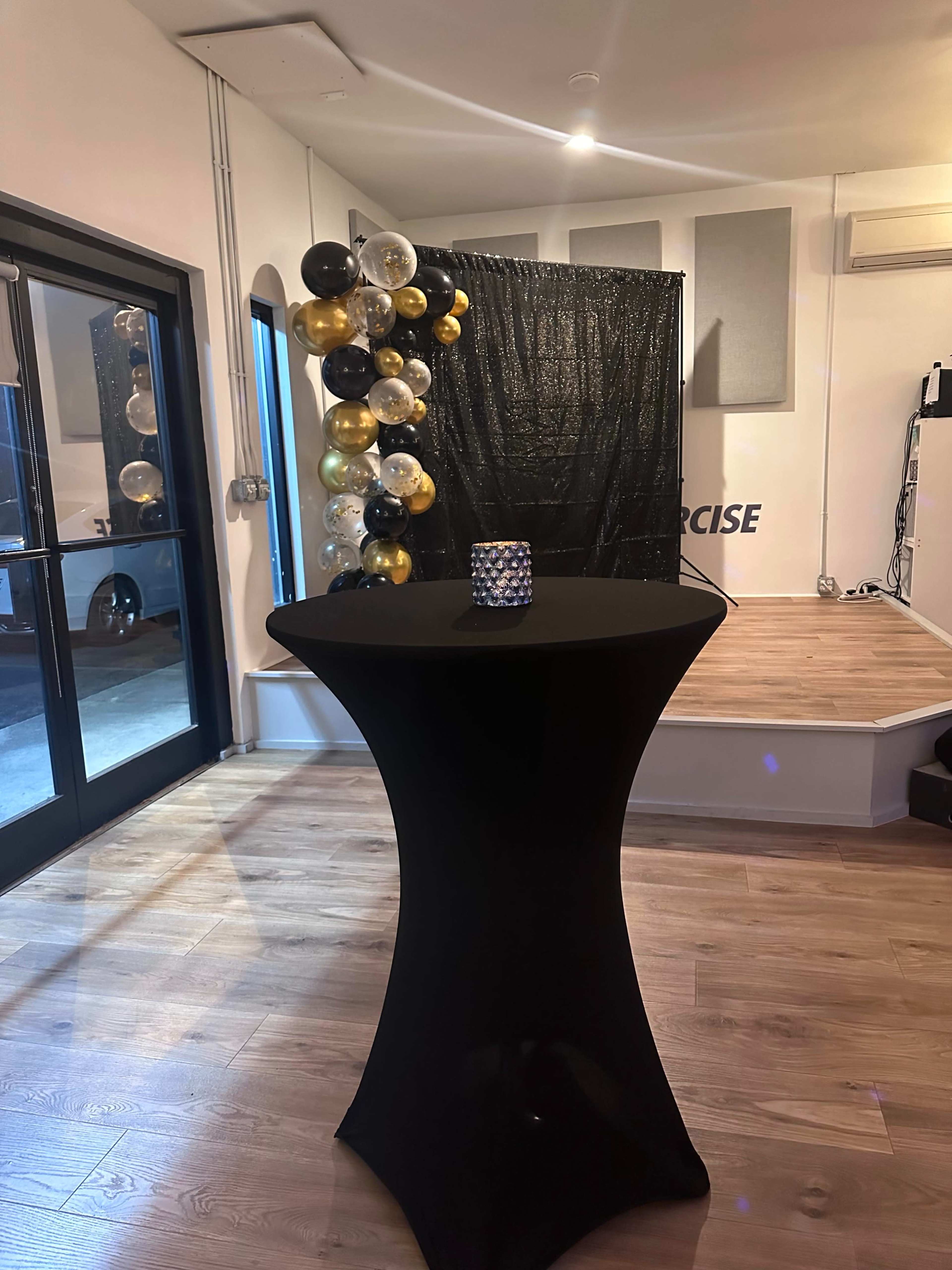 A black table is set in front of a backdrop adorned with black, gold, and clear balloons in a brightly lit room.